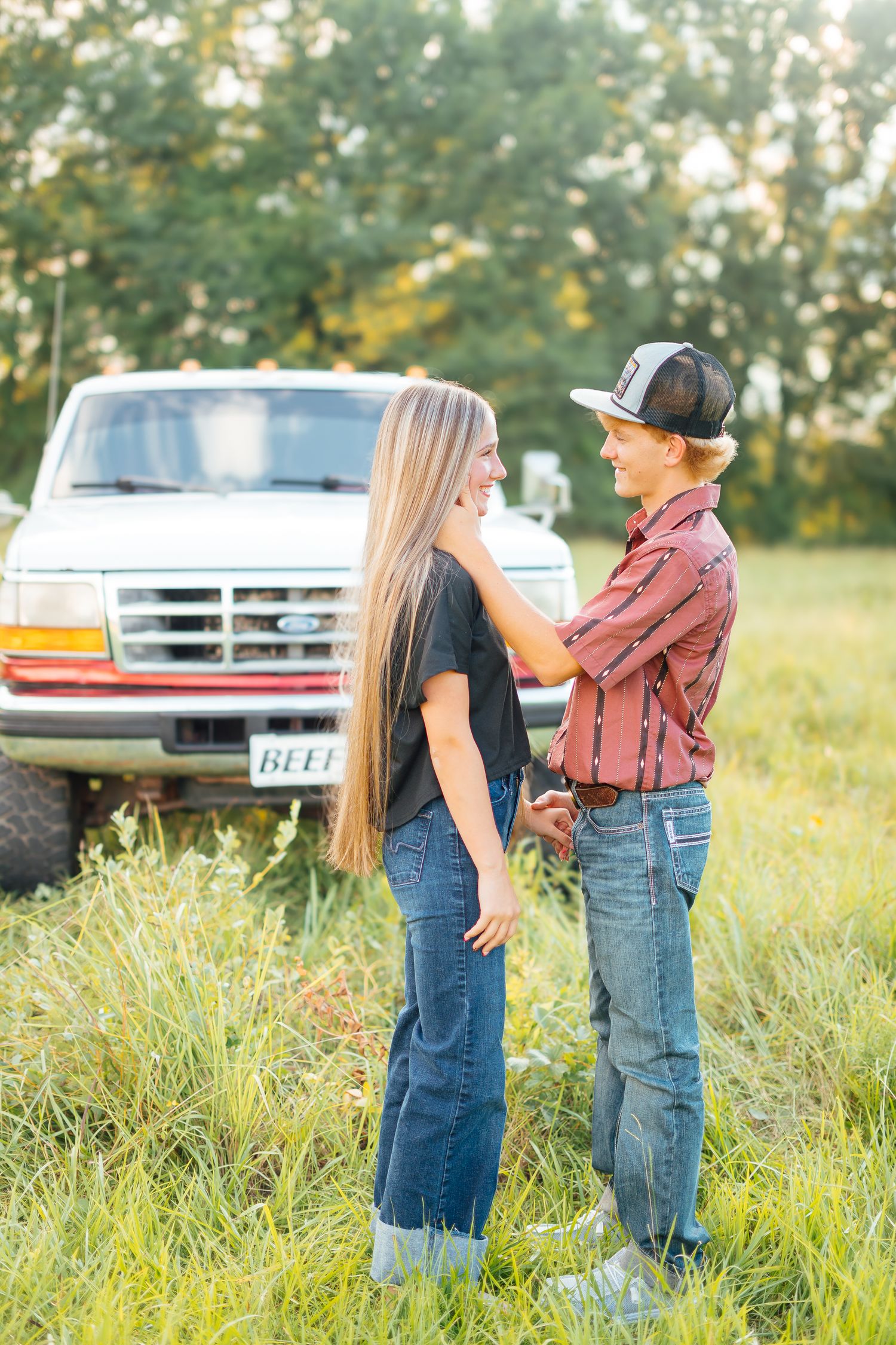 Young couple smiling and holding hands in a grassy field with a classic white pickup truck in the background.
