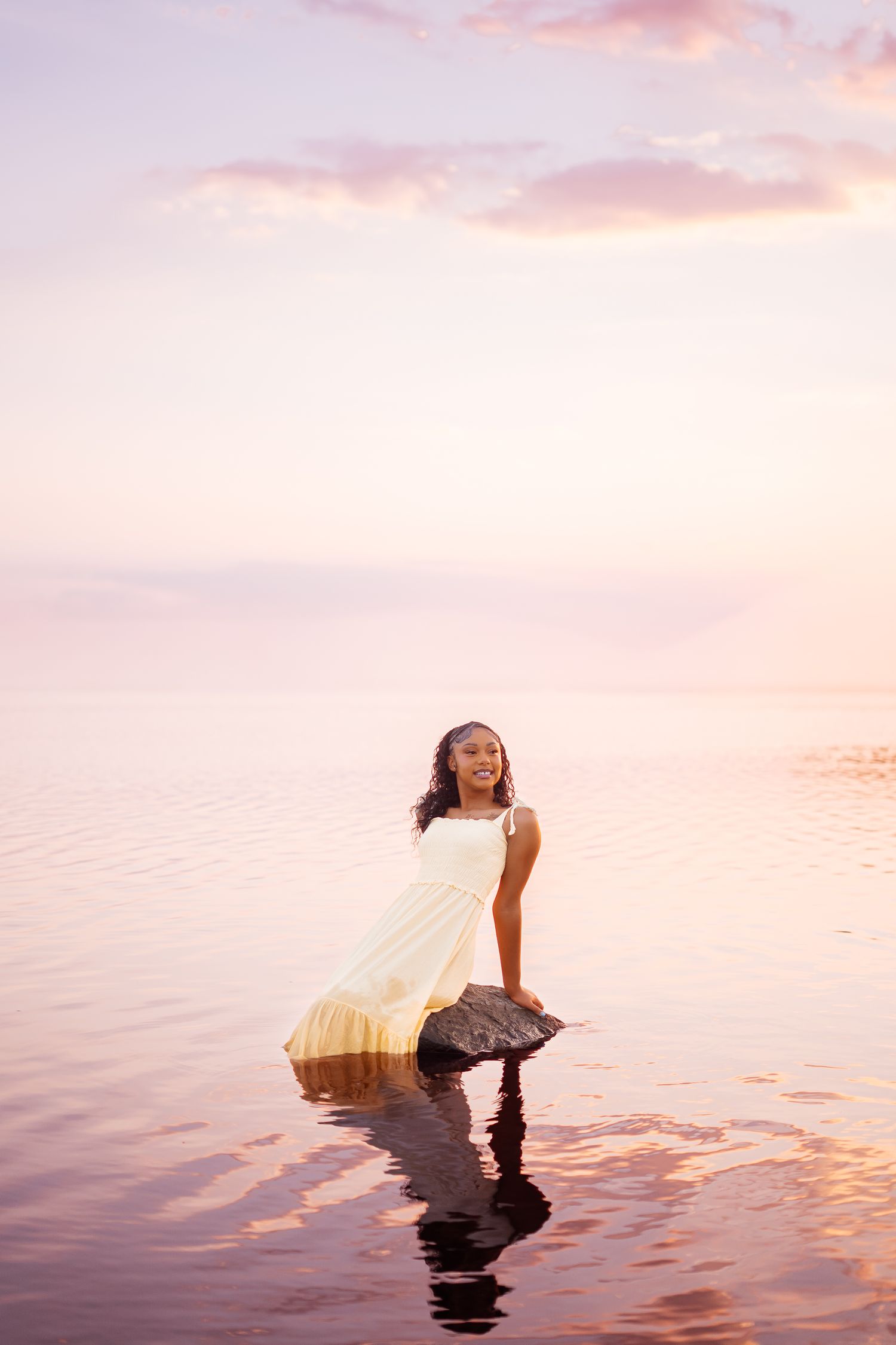 Young woman in a yellow dress stands on a rock in calm water during a pink and purple sunset.