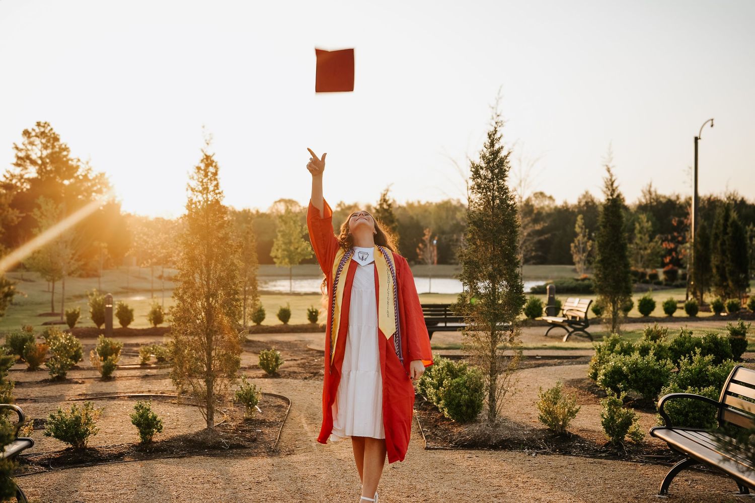 Graduate in red gown tosses cap in the air during golden hour outdoor graduation photo session.