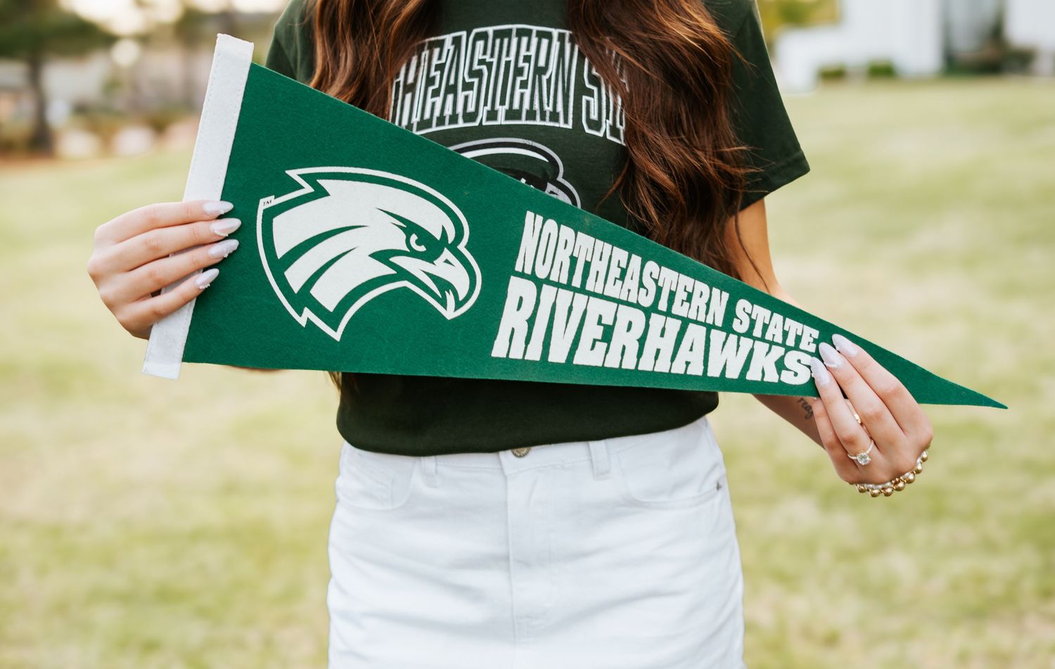 Young woman holding a green Northeastern State RiverHawks pennant flag outdoors on a sunny day.