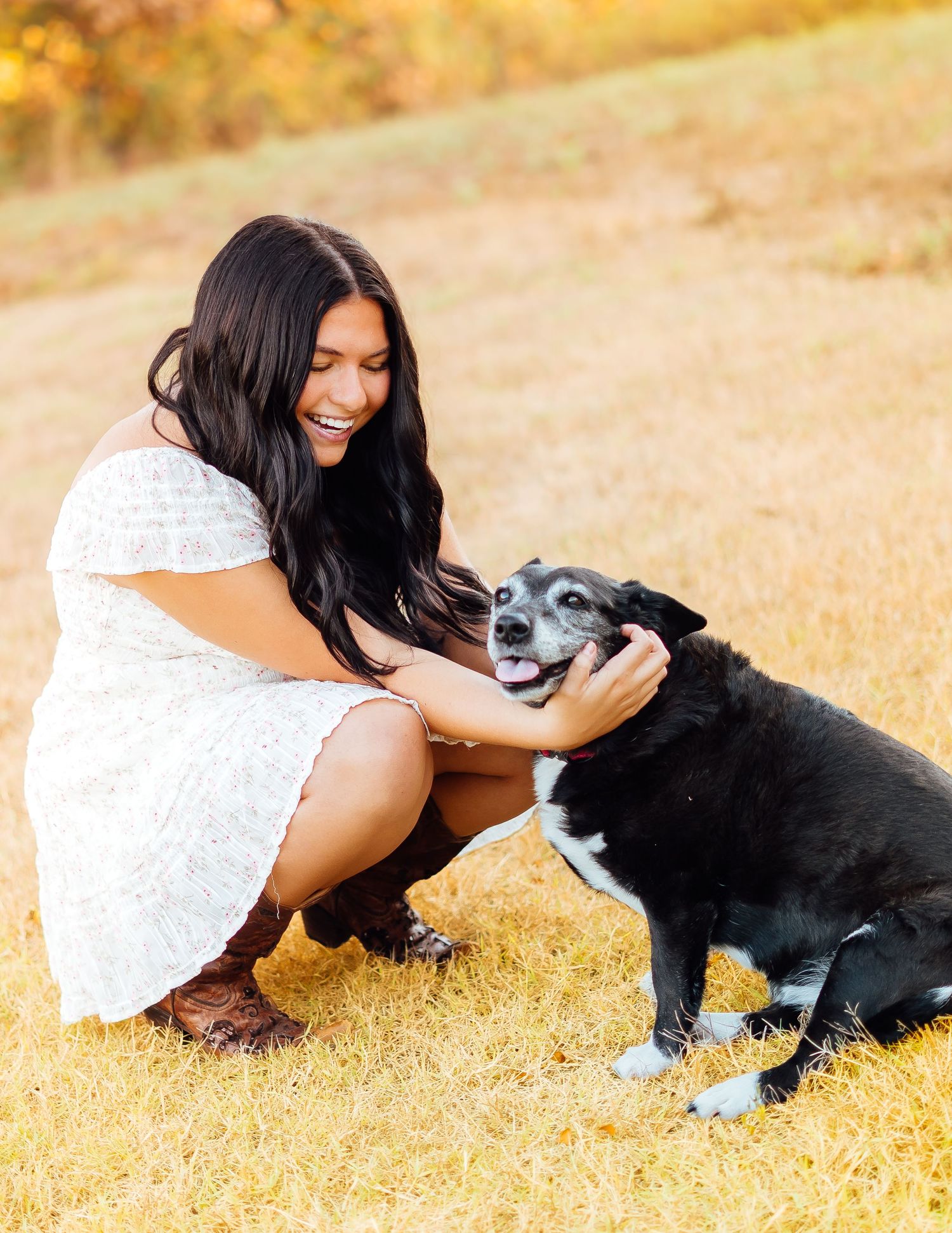 Young woman in white dress laughing while petting her black senior dog outdoors in golden light.