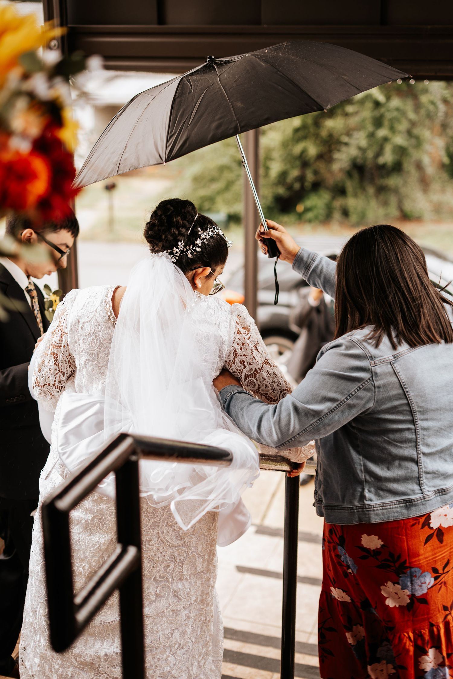 Wedding helper holding black umbrella over bride in lace gown and veil in the rain at outdoor venue.