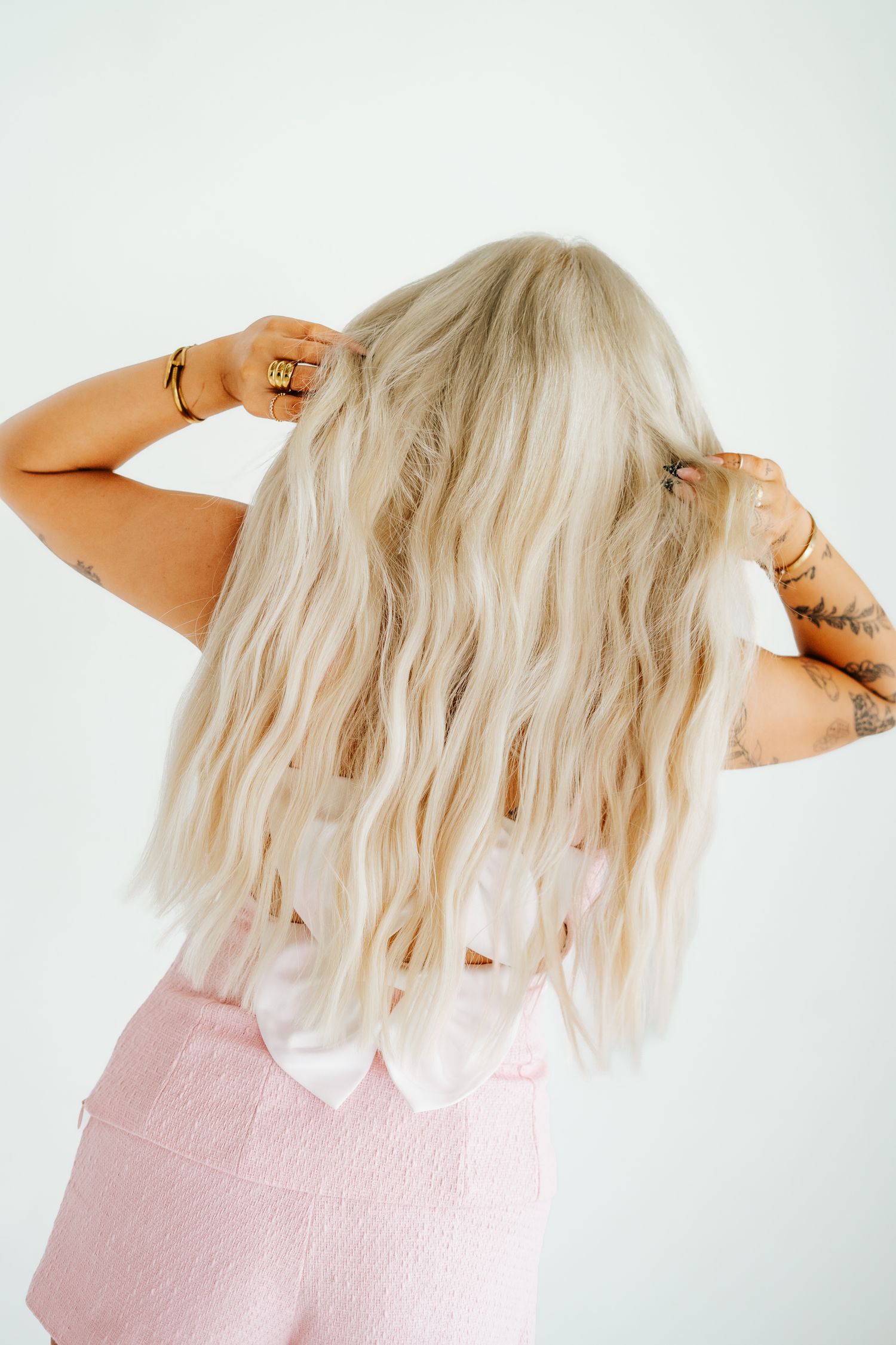 Back view of woman with long wavy blonde hair wearing pink skirt and gold jewelry.