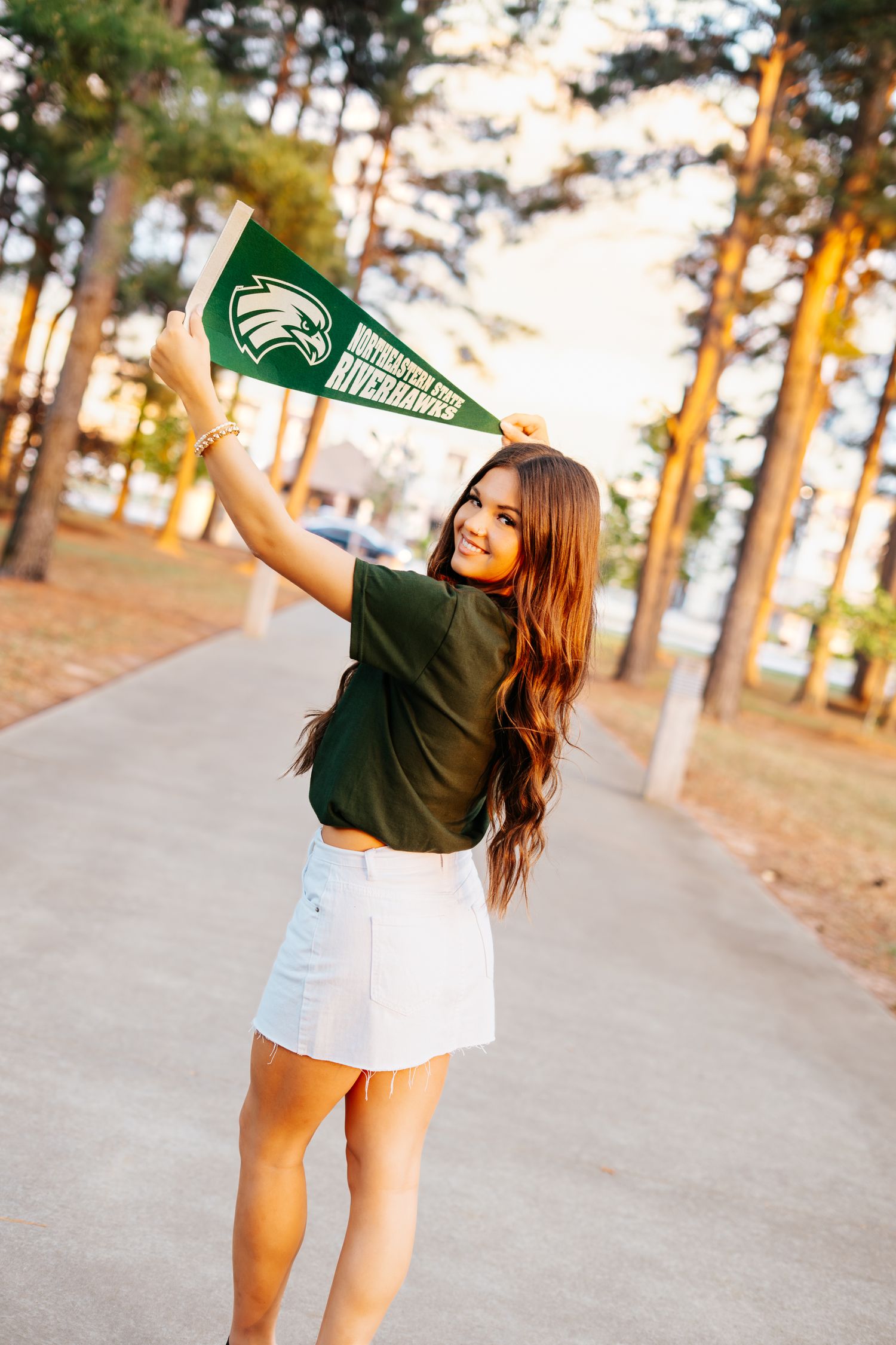 Smiling young woman holding green university pennant flag outdoors among tall pine trees.