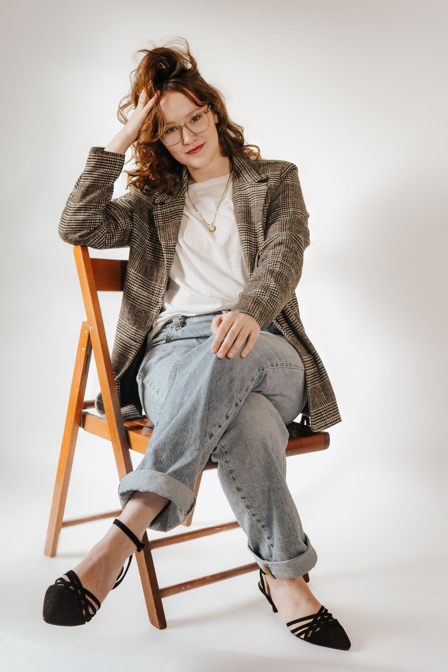 Redhead woman in plaid blazer and jeans sitting on a wooden chair against white background.