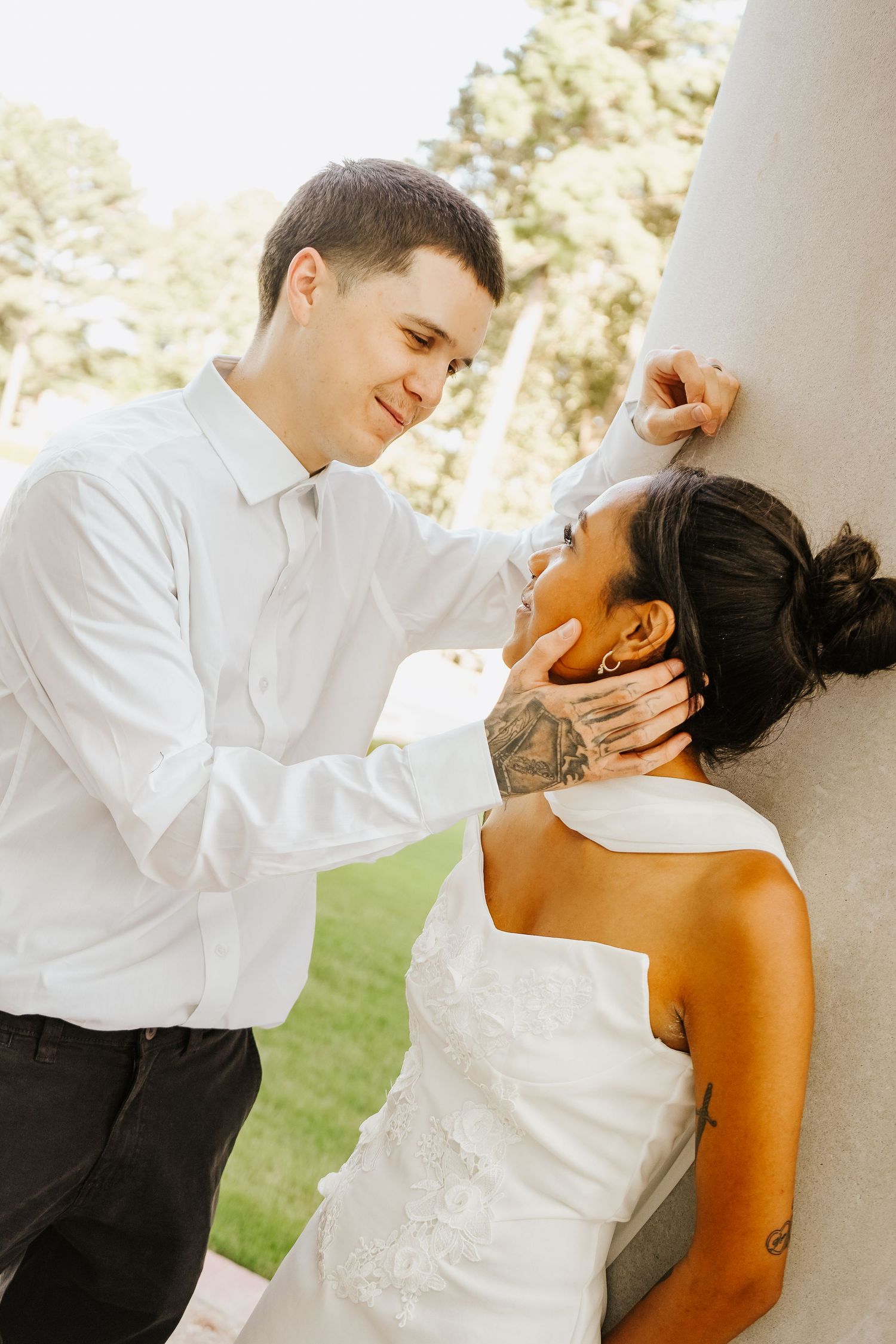 Groom in white shirt tenderly holding brides face near stone pillar outdoors at intimate wedding portrait session.