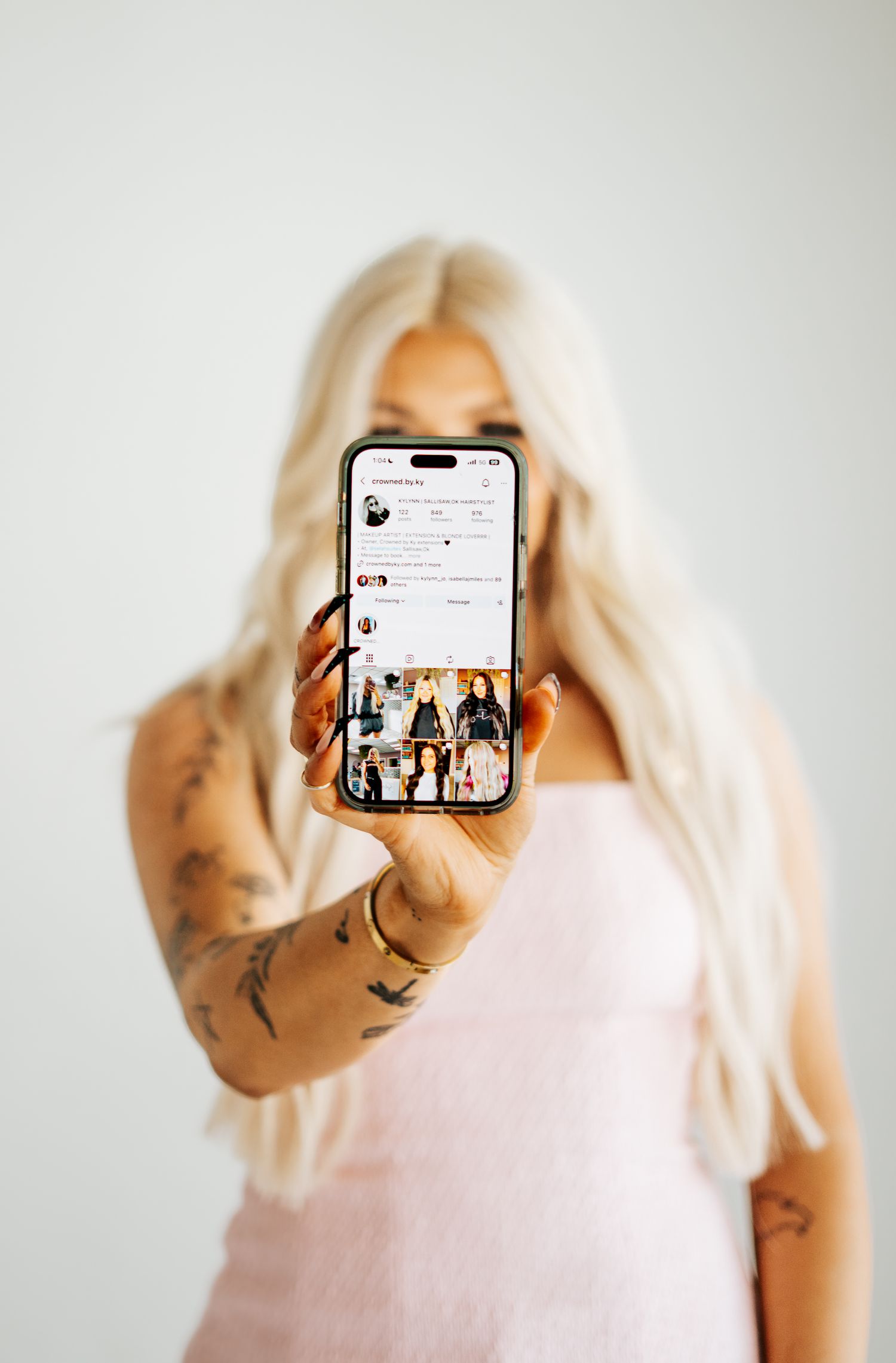Tattooed blonde woman holding up smartphone displaying a fashion Instagram profile page.