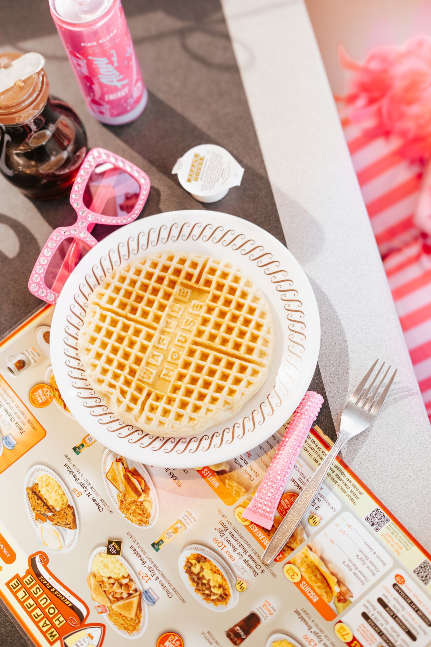 Overhead view of a waffle on a plate with pink sunglasses and Waffle House menu on table.