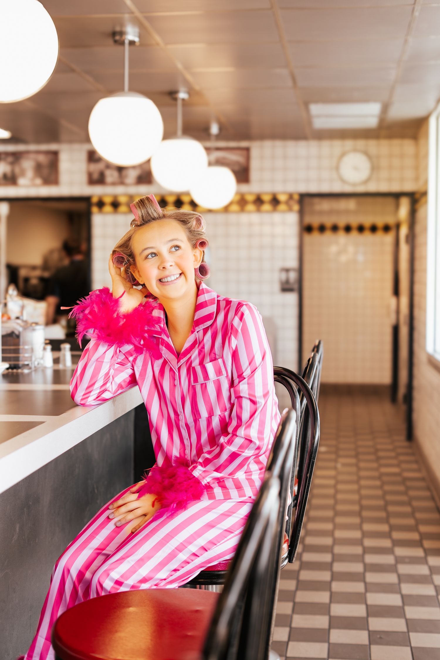 Smiling woman in pink striped pajamas with feather boa sitting at a retro diner counter.