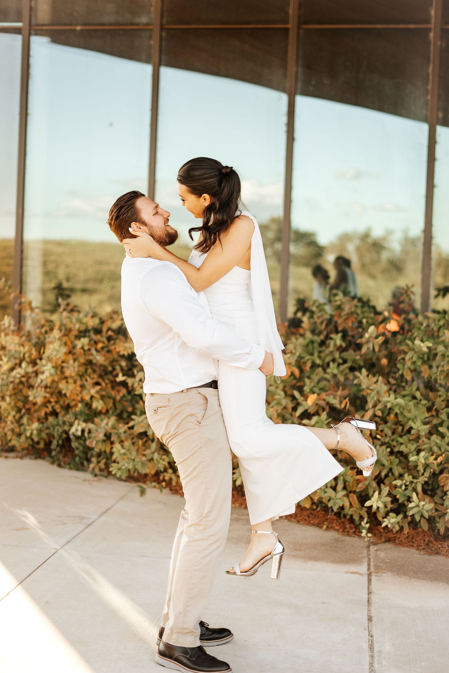 Groom lifting bride in white strapless wedding dress in front of reflective glass building exterior.