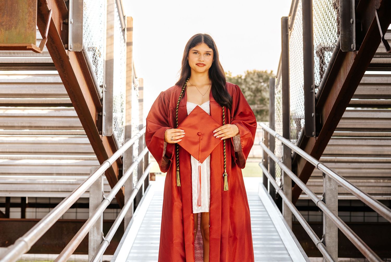 High school graduate in red cap and gown holds diploma on a metal bridge structure outdoors.