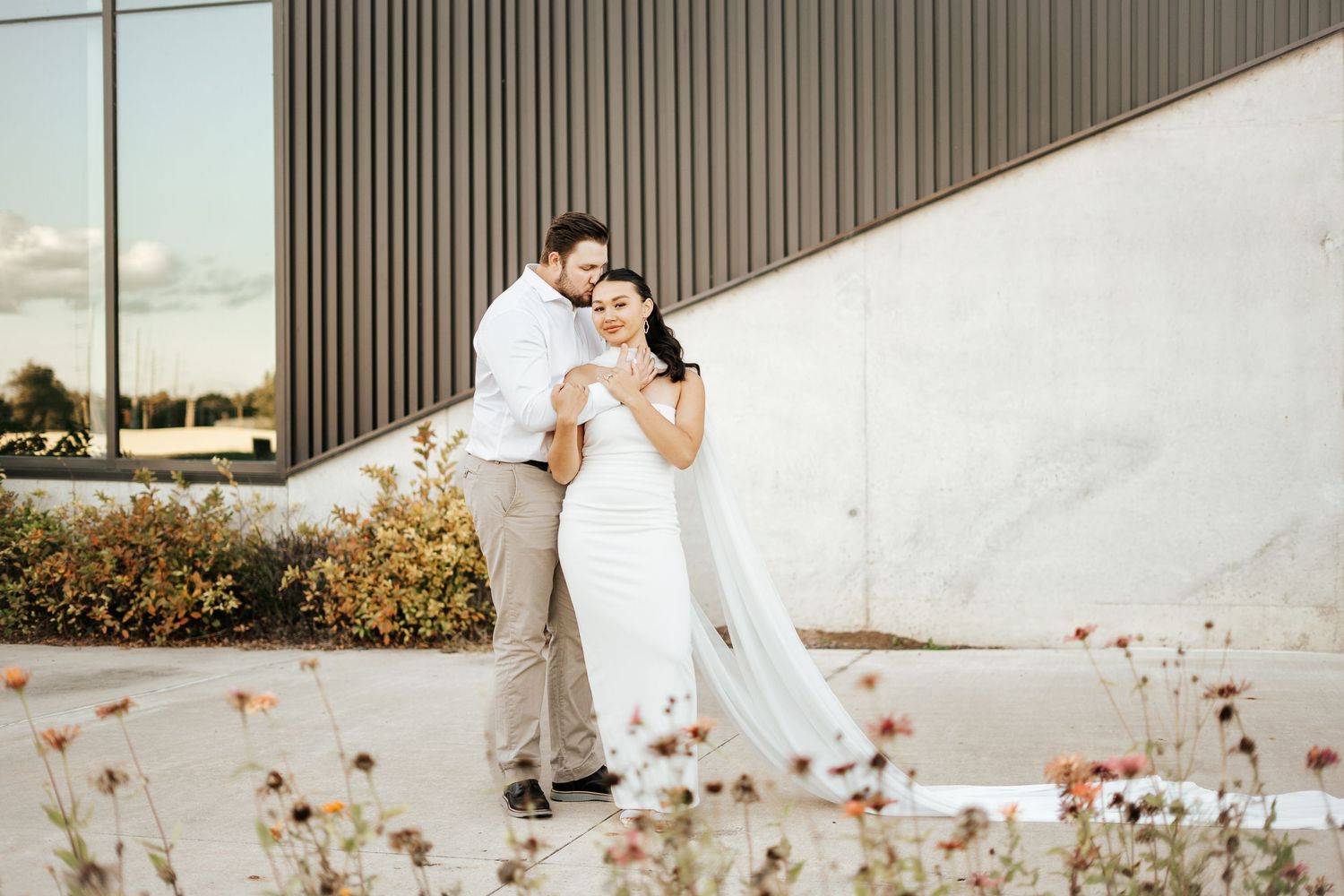 Groom embracing bride in white wedding dress outdoors near modern building with wildflowers in foreground.