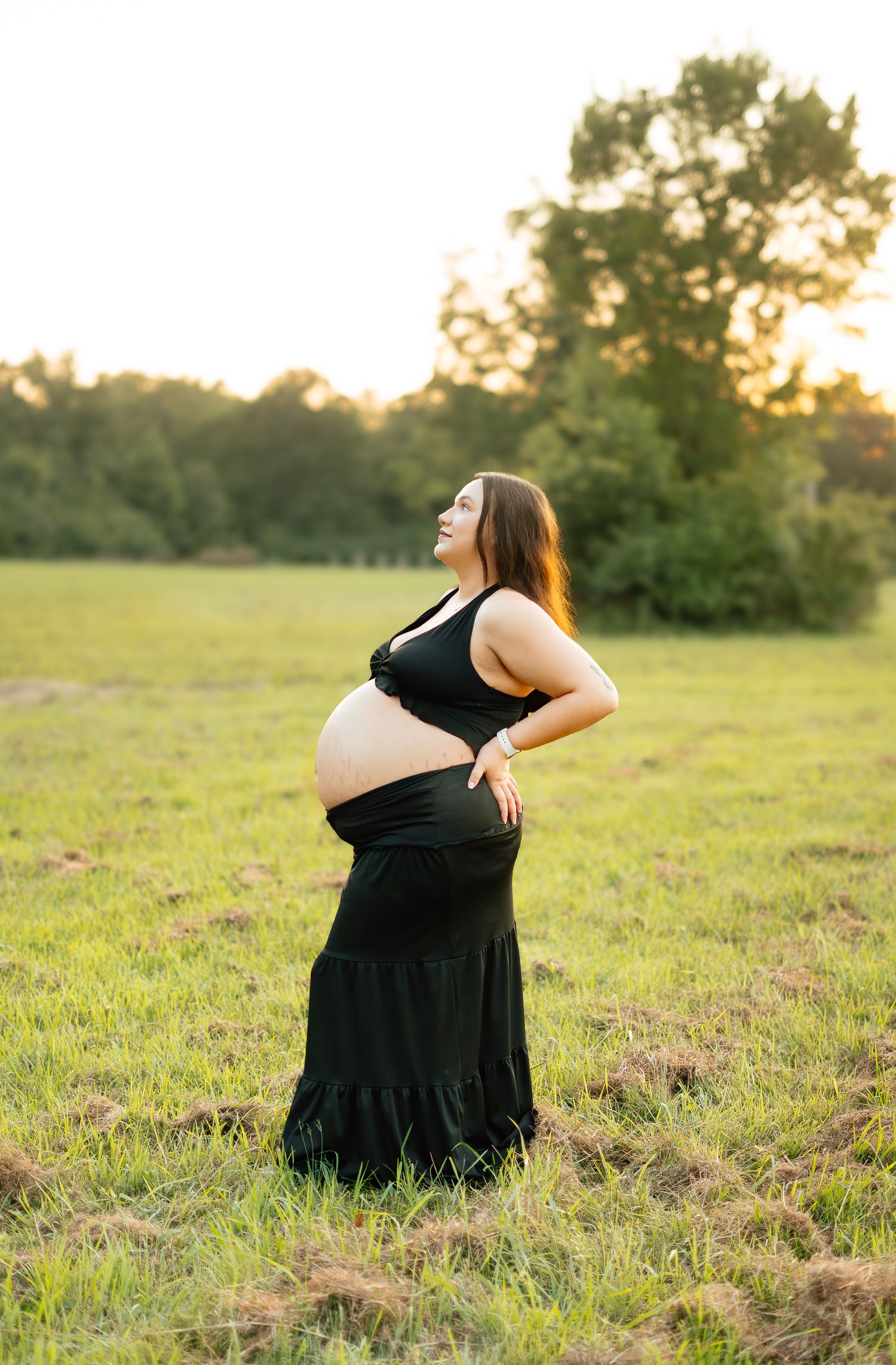 Pregnant woman in black crop top and skirt posing in green field at sunset.