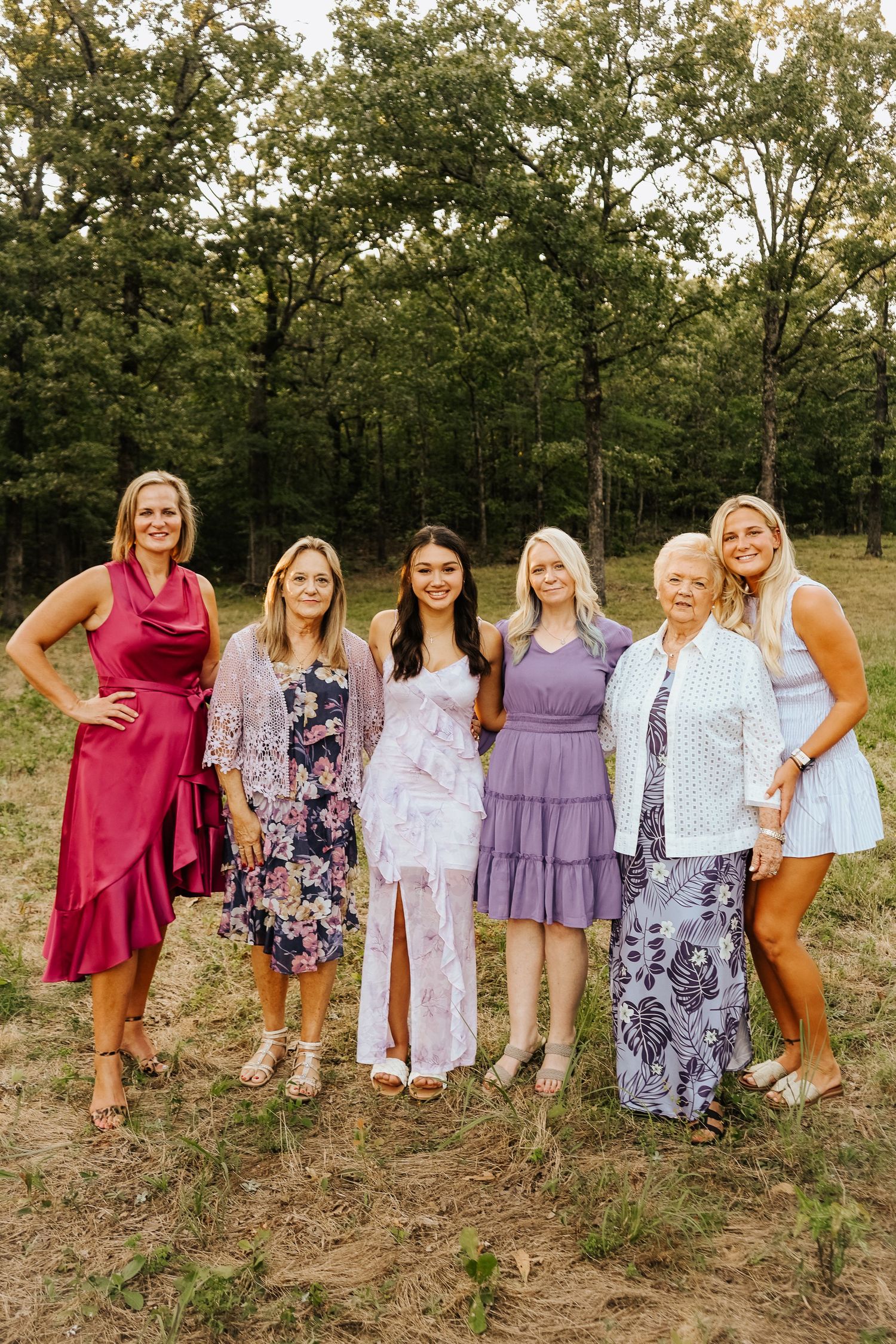 Group of six women in colorful dresses posing together outdoors near trees.