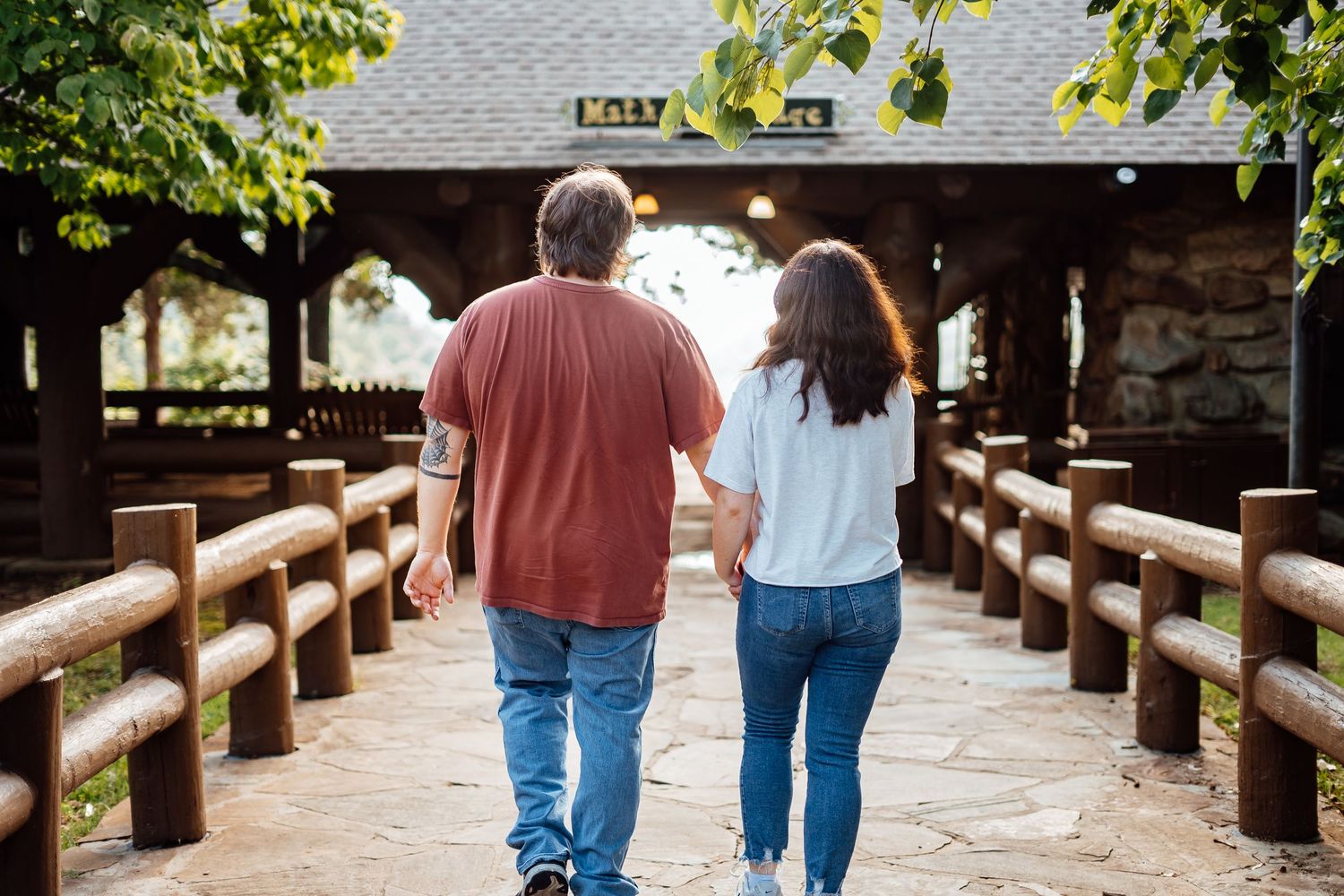 Couple walks hand in hand through a rustic wooden covered bridge pathway on a sunny day.