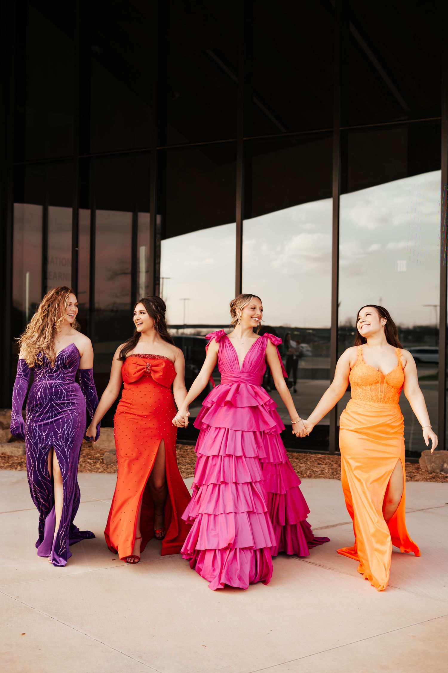 Four women in vibrant purple, orange, pink, and orange formal gowns holding hands outside a building.