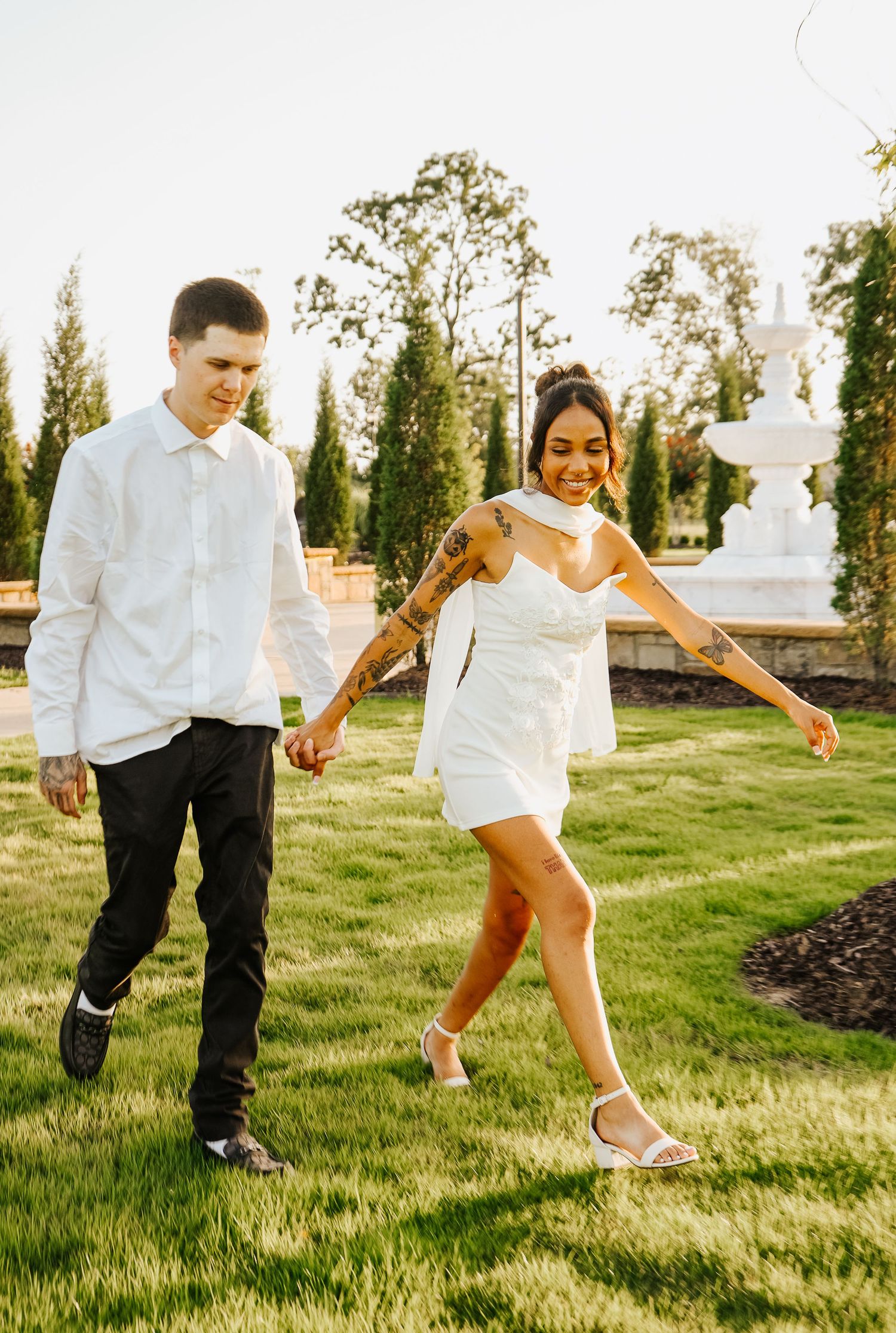 Happy couple walking on green lawn during golden hour, woman in white mini dress laughing with tattoos.