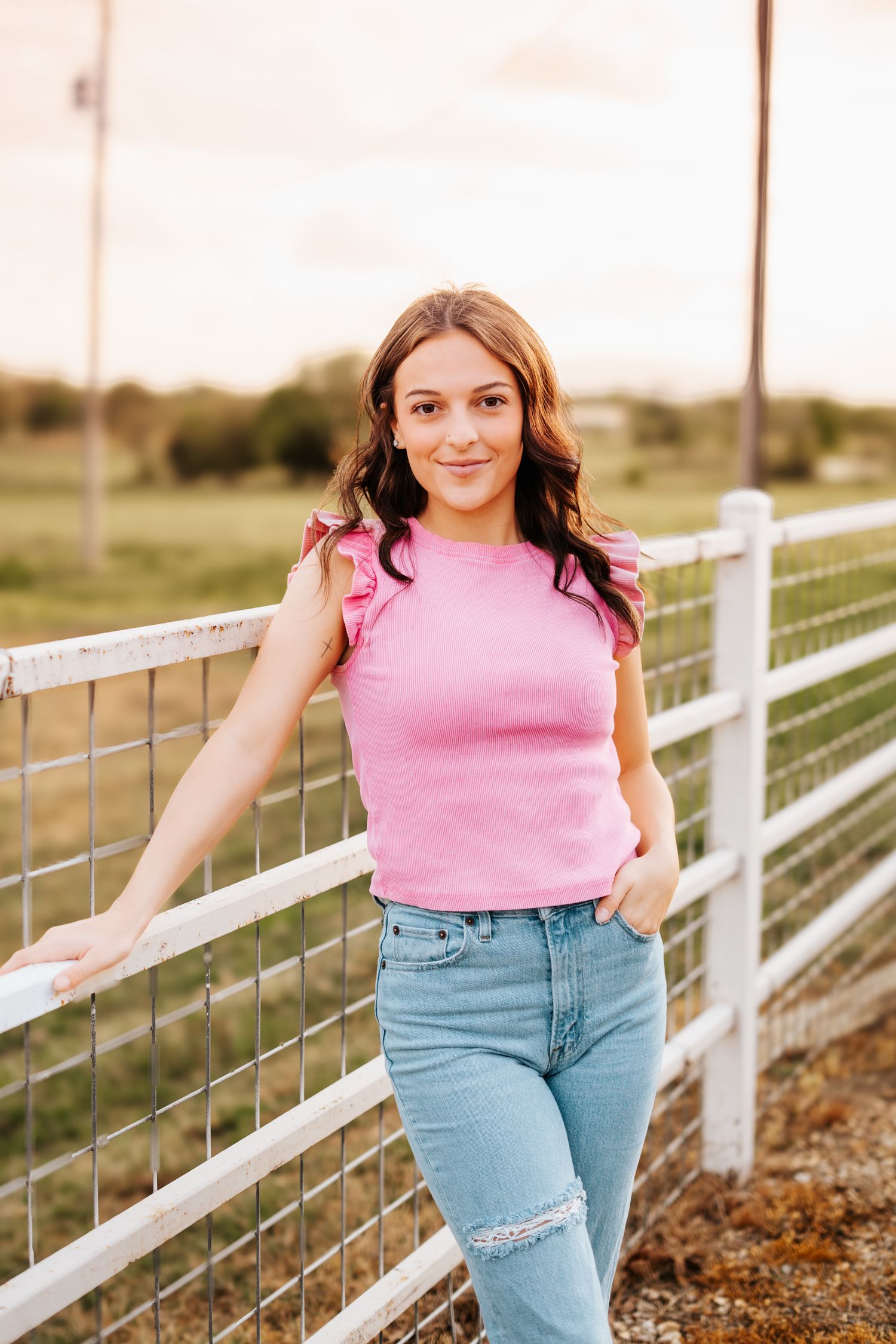 Teen girl in a pink ruffled top and ripped jeans leans against a white fence at golden hour.