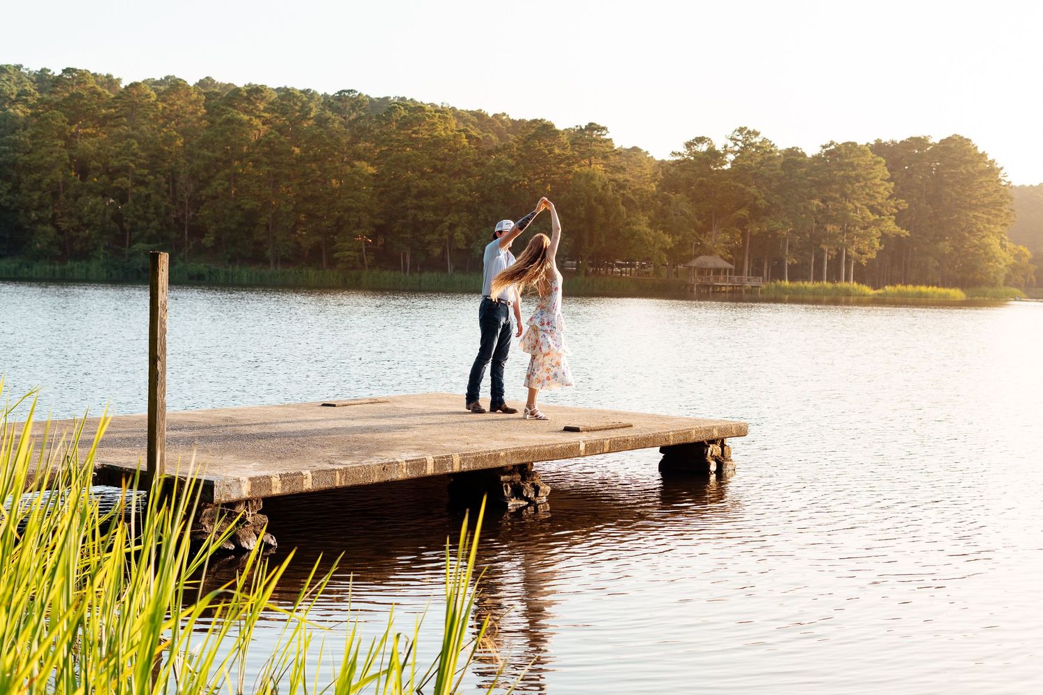 Romantic couple dancing on a wooden dock by a serene lake surrounded by lush green trees at sunset.
