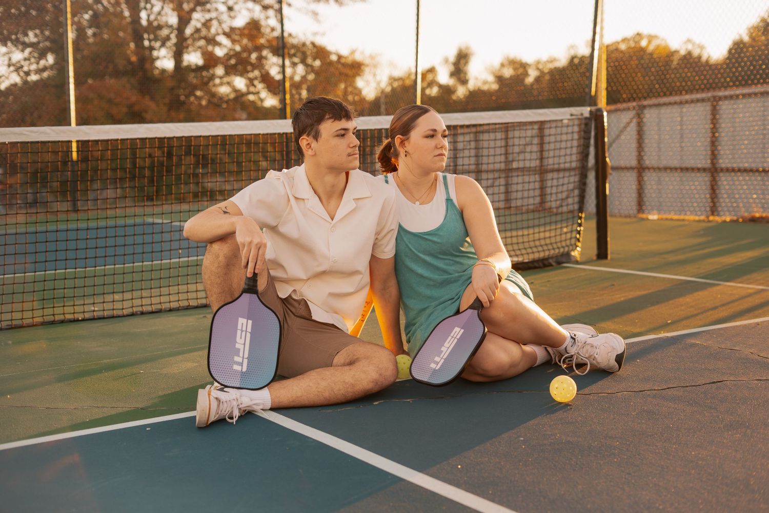 Couple sitting back-to-back on a pickleball court holding paddles during golden hour.
