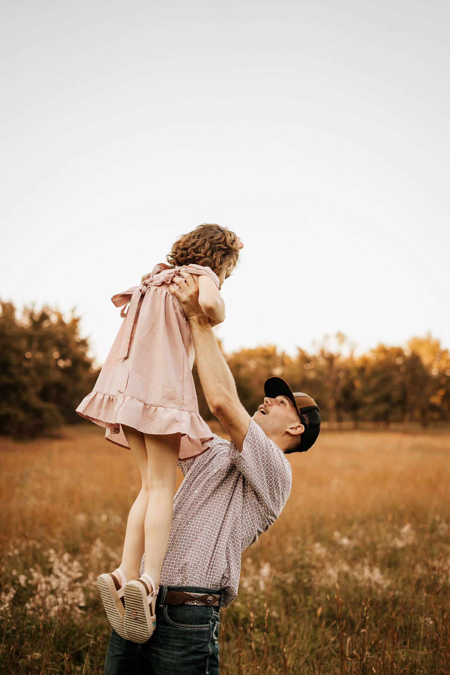 Father lifting young daughter in pink dress above his head in a golden autumn field.