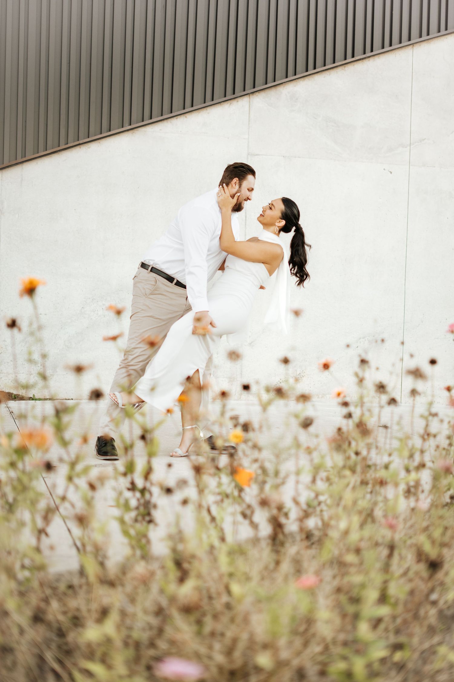 Groom dipping bride in white wedding dress among wildflowers against a modern concrete wall backdrop.