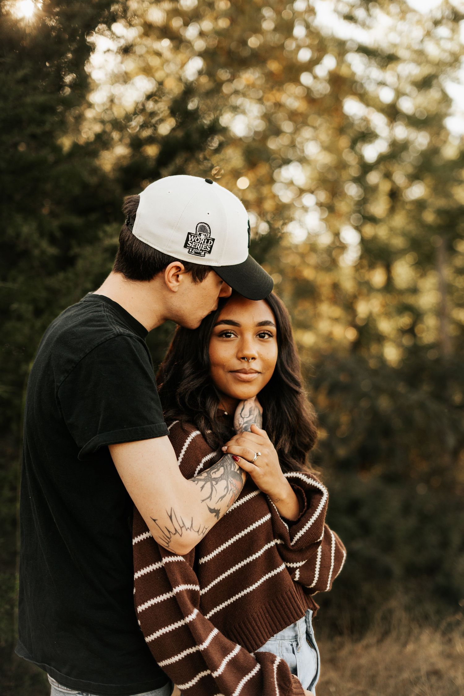 Young couple embraces in golden sunset light among pine trees, woman in striped brown sweater.