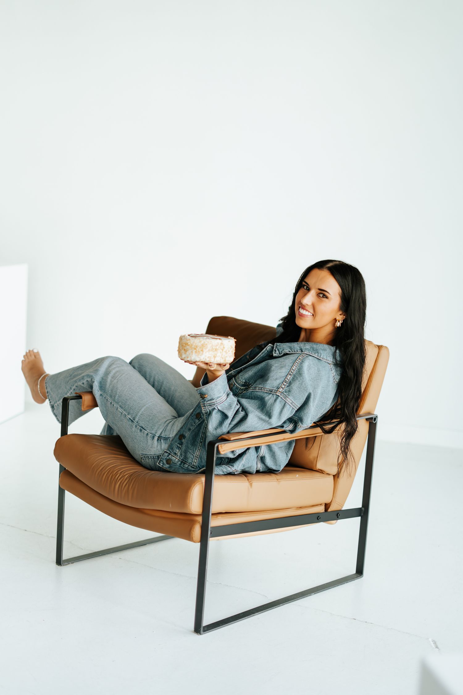 Brunette woman relaxing in brown leather chair holding a book in a bright white studio.
