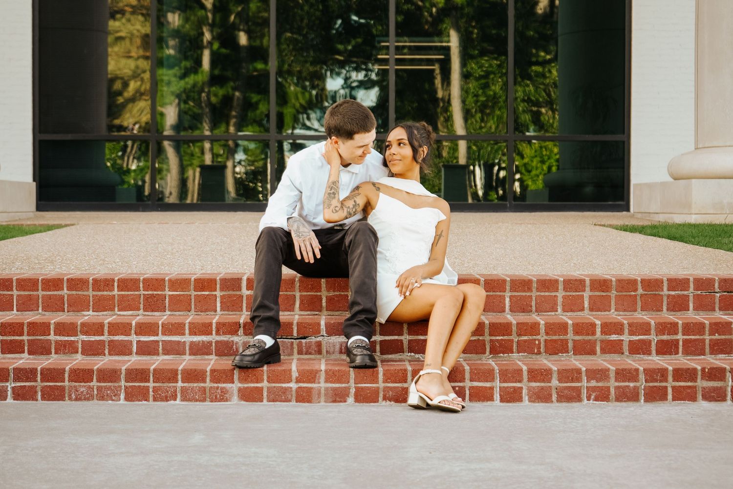Couple sitting on brick steps during engagement session, man in dress shirt and woman in white mini dress.