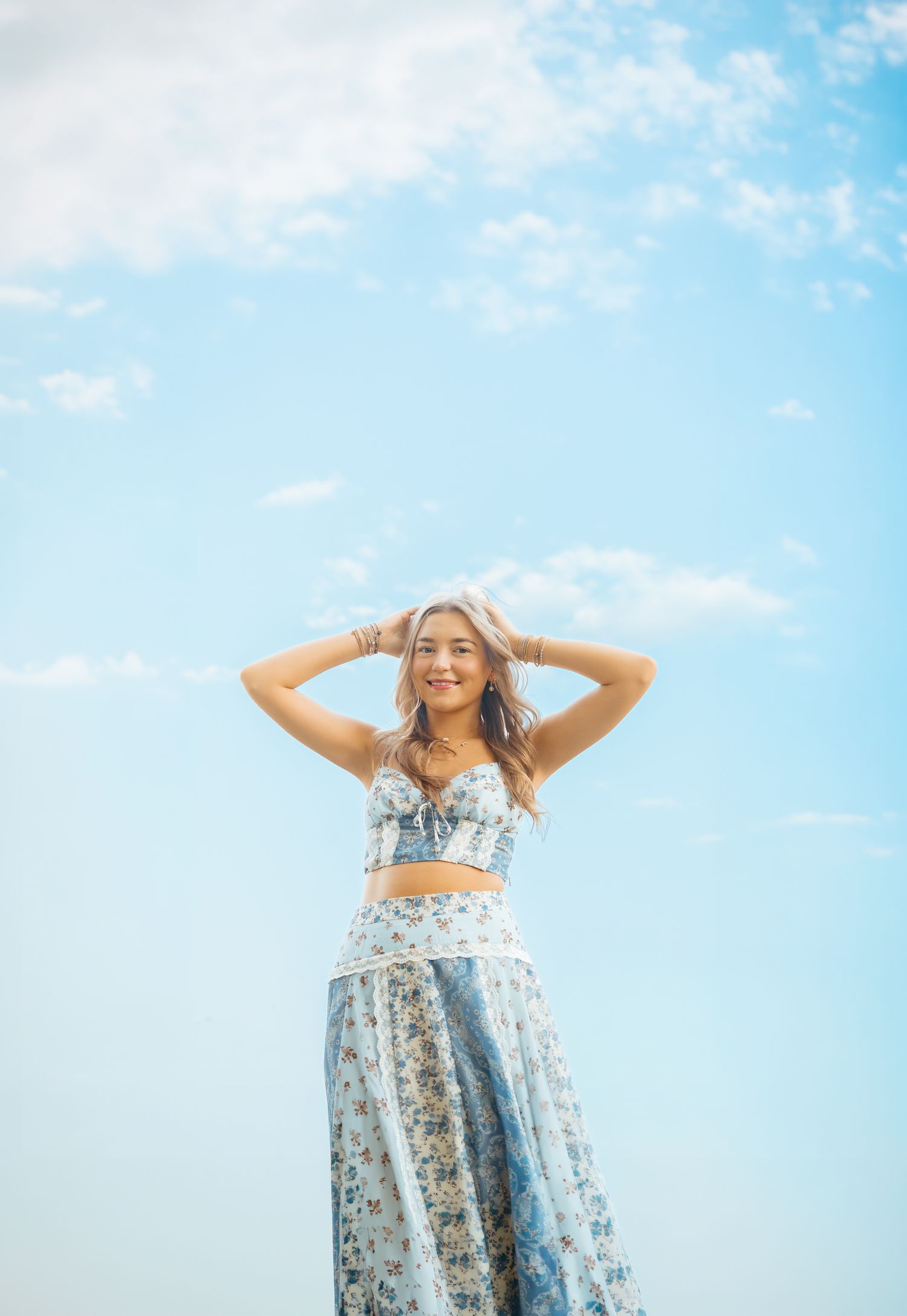 Young woman in a floral blue crop top and maxi skirt poses with hands in hair against a blue sky.