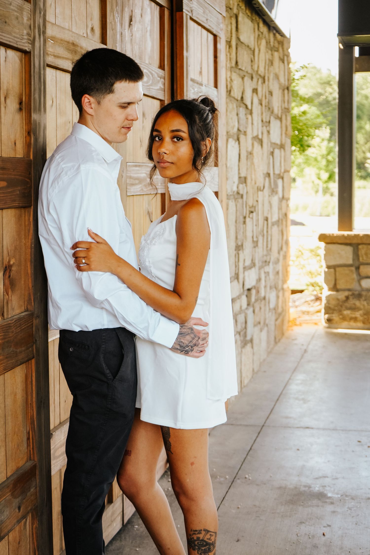Couple embracing by rustic wooden doors, woman in white halter mini dress with tattoos leaning against man.