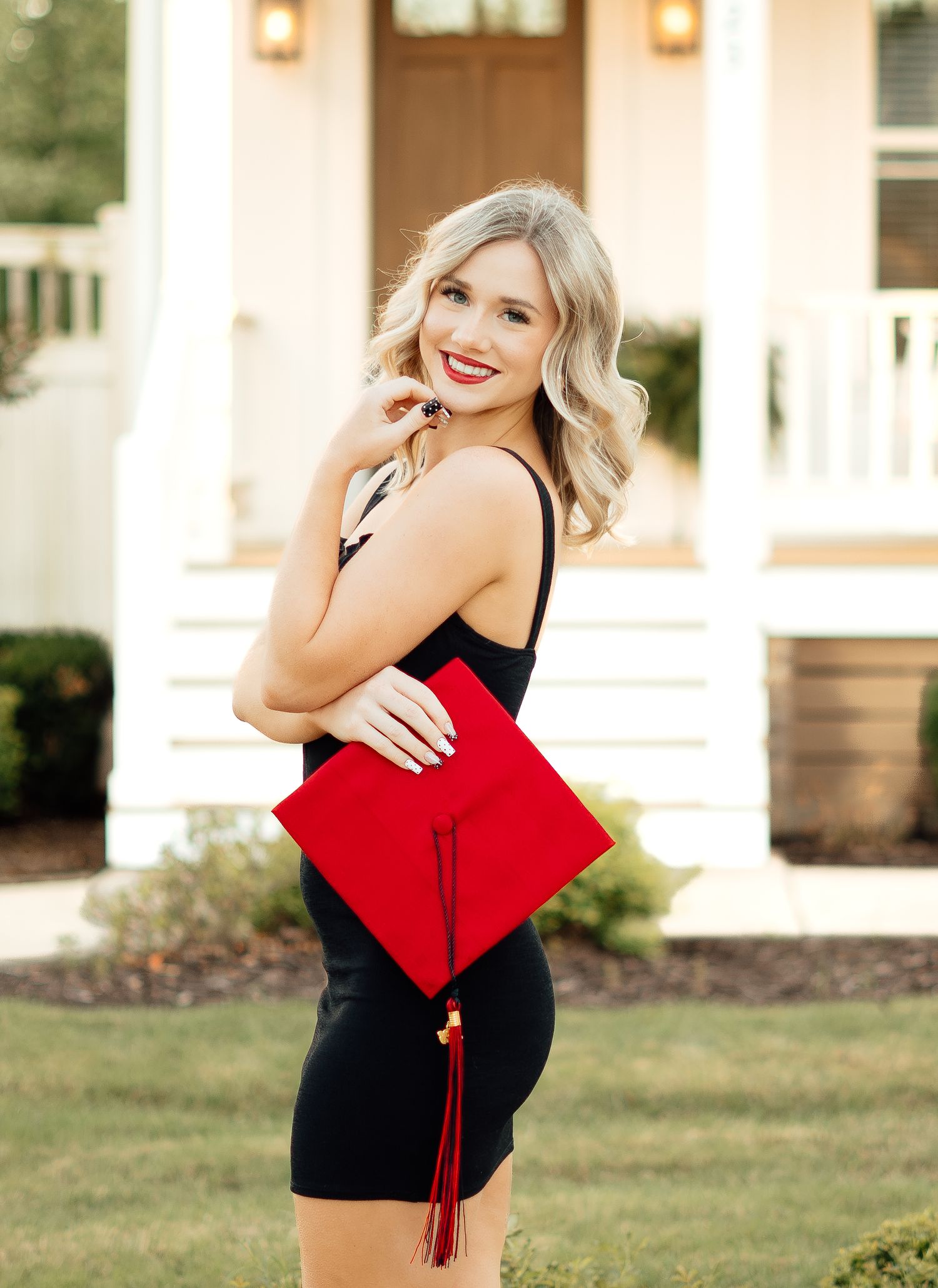 Blonde woman in black dress holding red graduation cap with tassel in front of a white house.