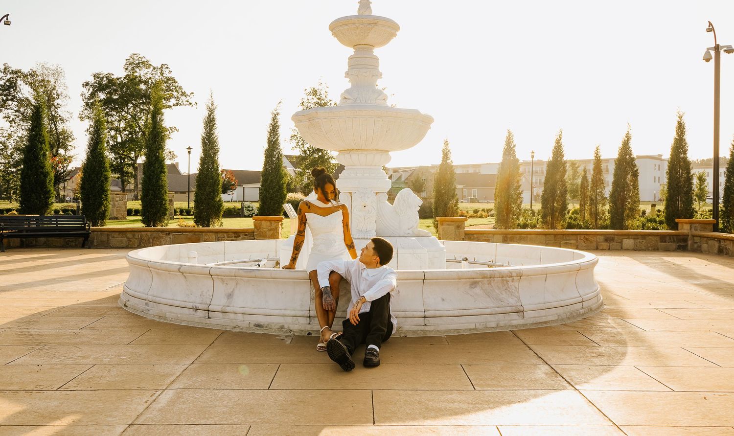 Couple at white marble fountain during golden hour engagement session, man kneeling before woman in white dress.