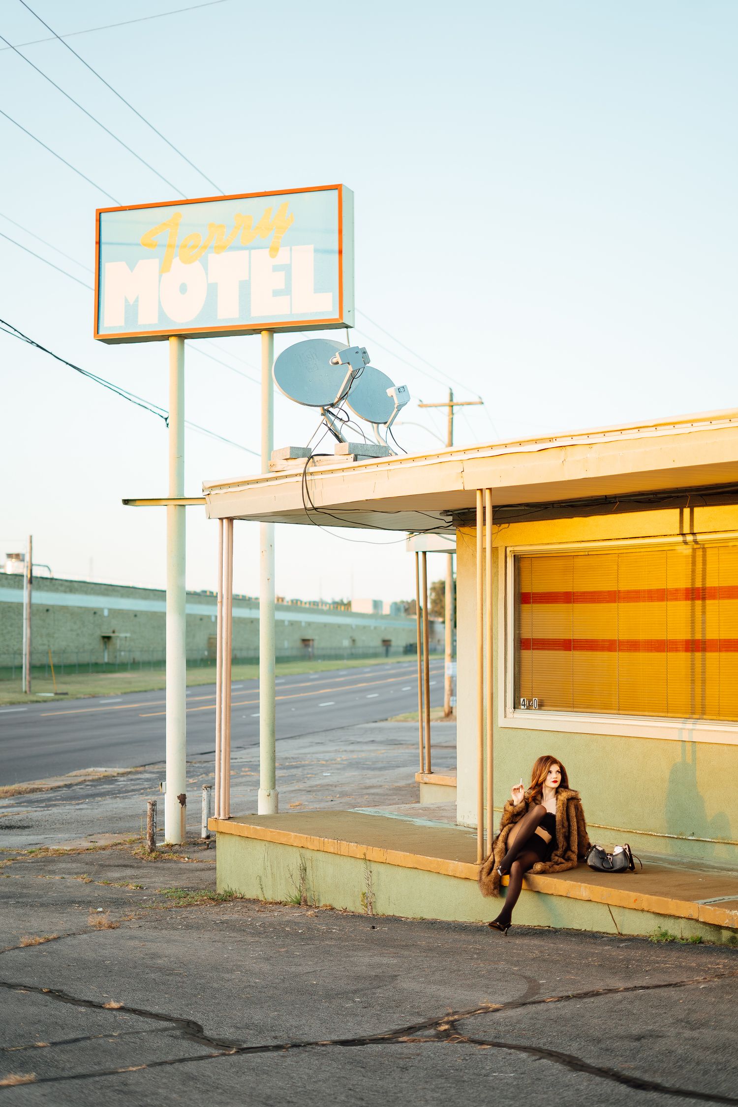 Woman in fur coat sitting outside a retro motel at golden hour on a quiet highway.