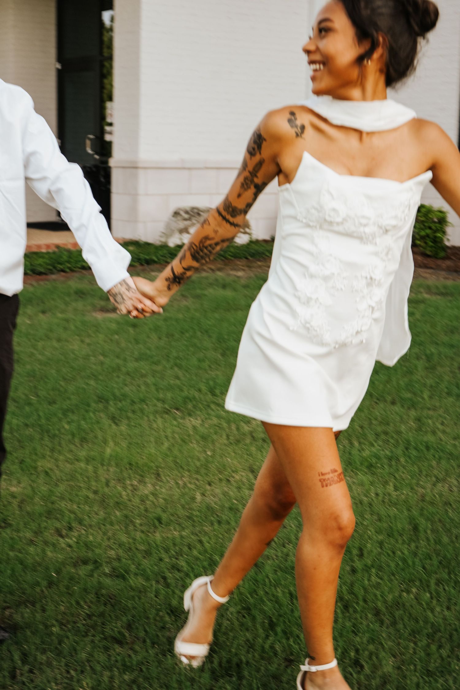 Tattooed bride in short white halter wedding dress holding grooms hand and laughing while walking on green lawn.