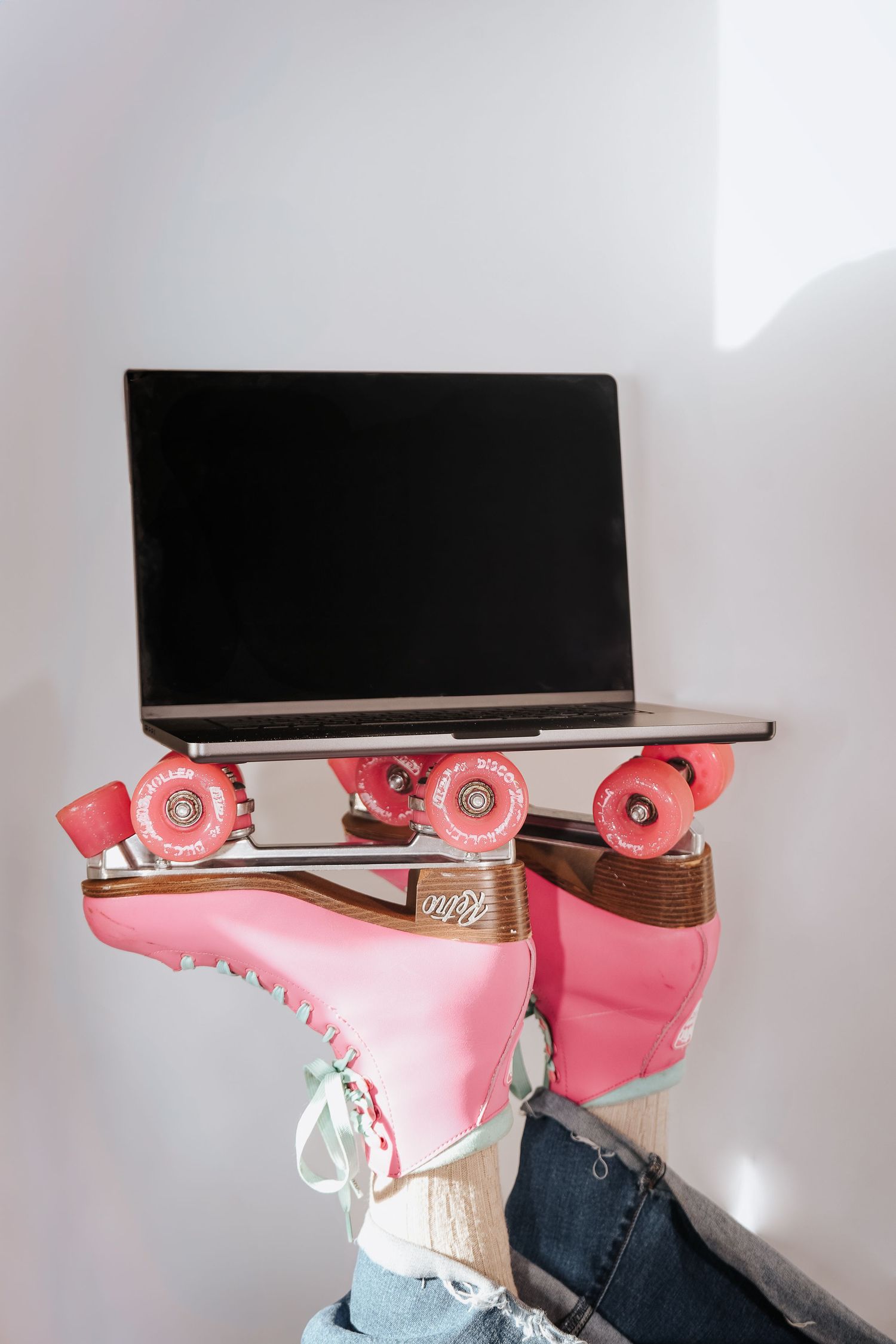 Person wearing pink roller skates balancing a laptop on their feet against a white wall.