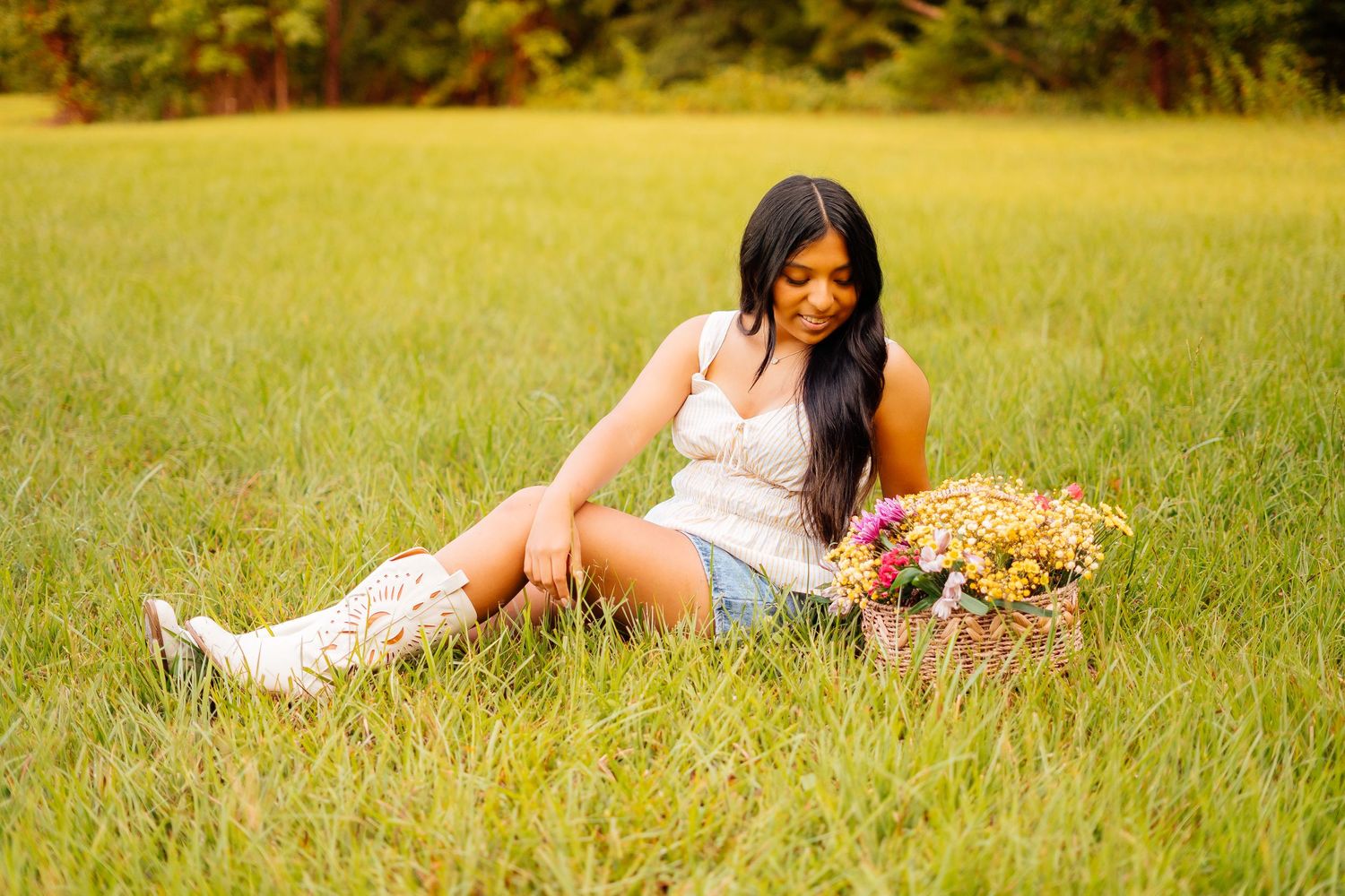 Young woman in white lace top and denim shorts sits in green grass with a flower basket beside her.
