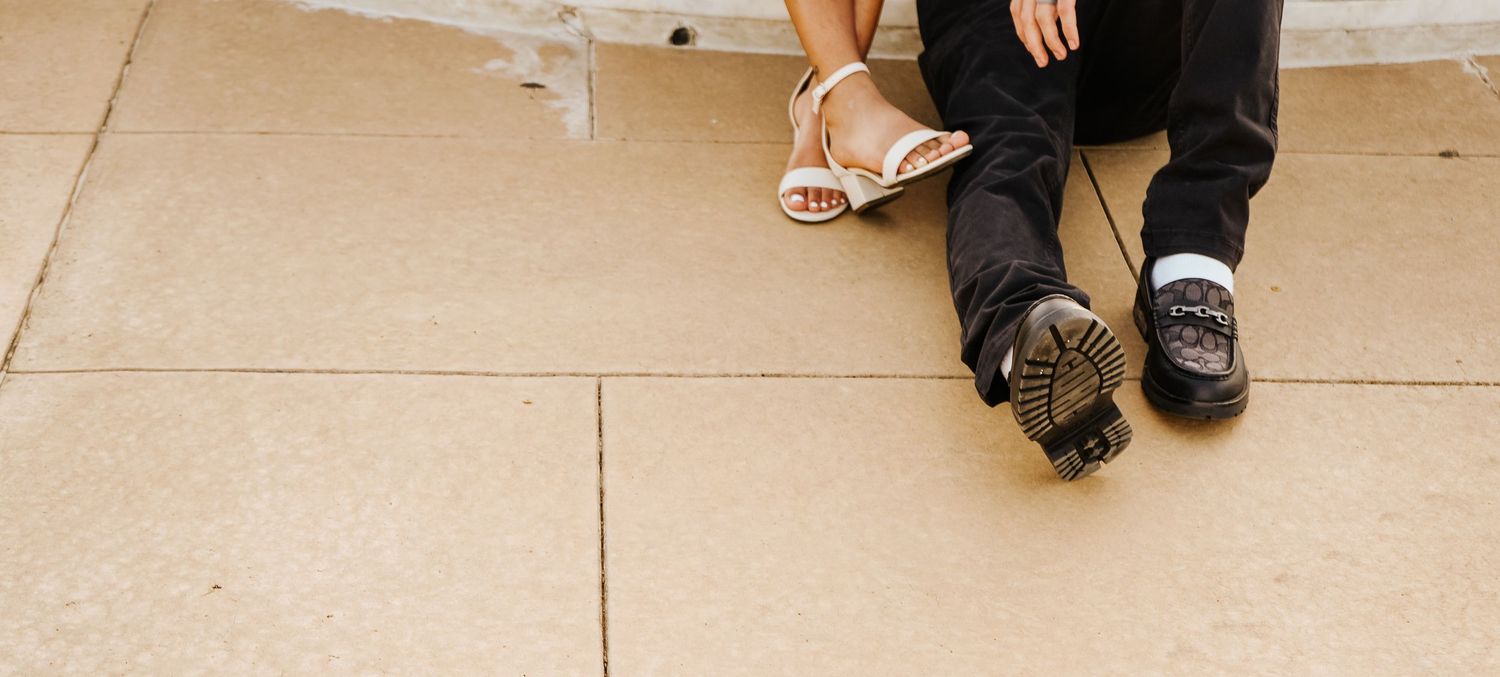 Close-up of couples feet sitting together, woman in strappy heels and man in dark loafers on pavement.