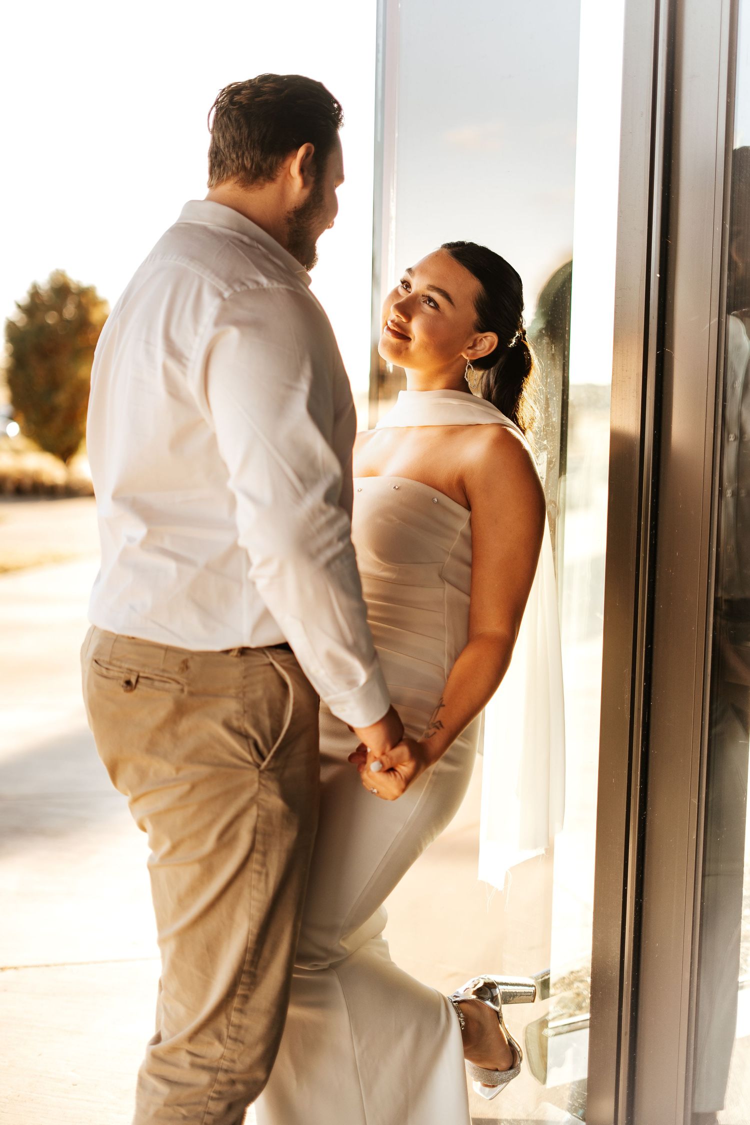 Bride in a white halter gown smiling at groom in white shirt holding hands near a sunlit glass door.
