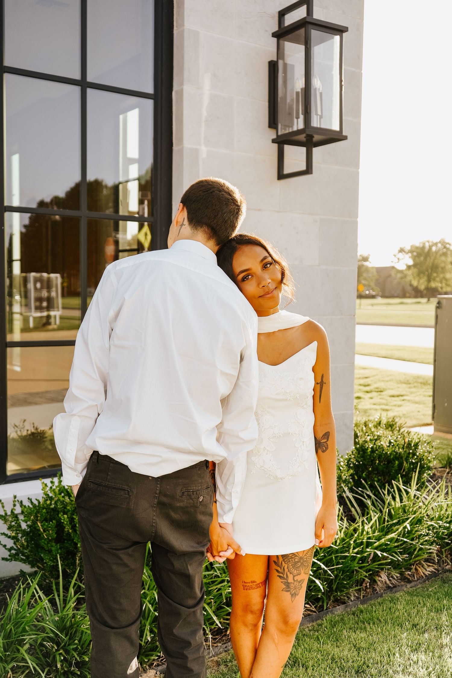 Woman in white mini dress with tattoos resting head on mans shoulder during golden hour engagement photos.