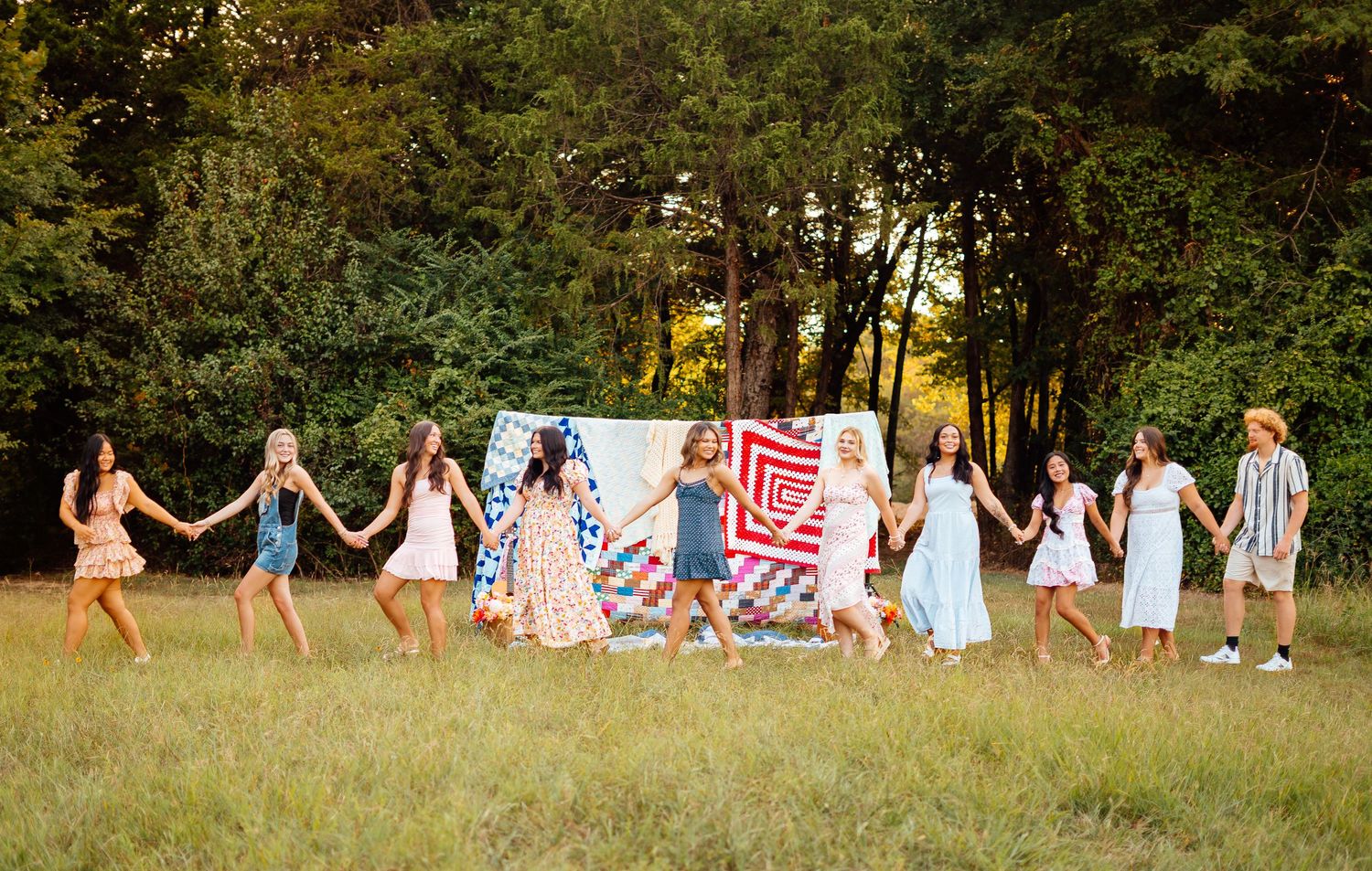 Group of young women holding hands in a line outdoors in a sunny field with colorful quilts displayed behind them.