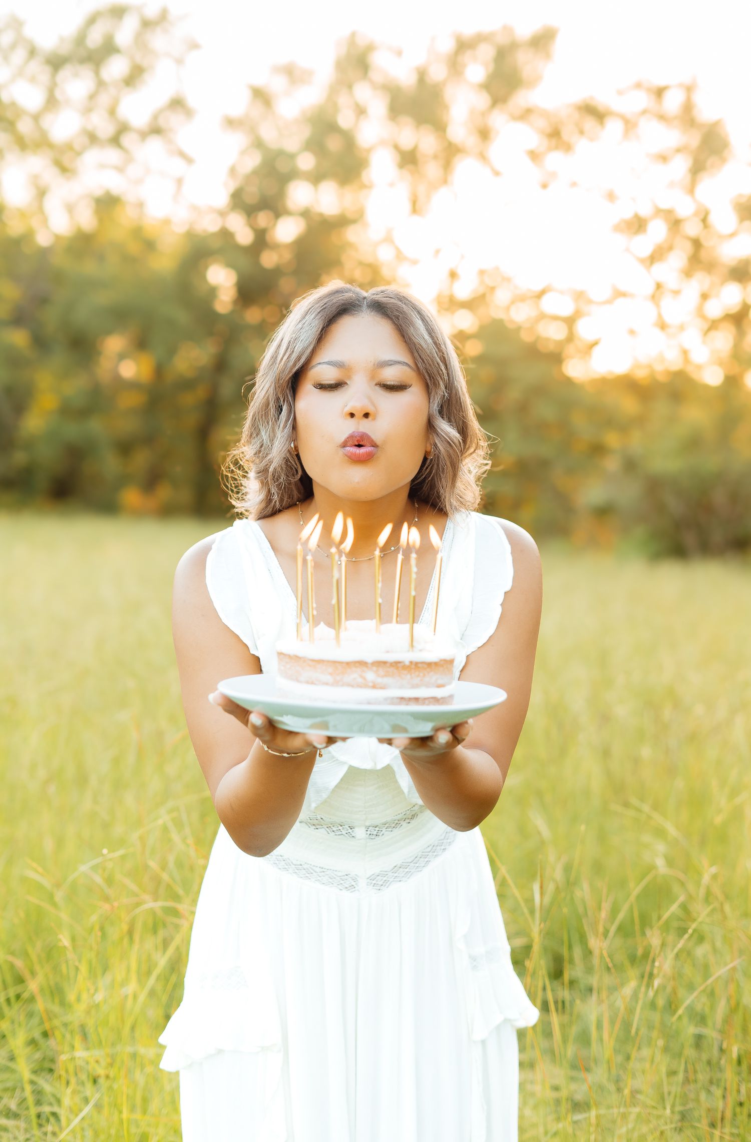 Young woman in white dress blows out candles on birthday cake in golden sunlit meadow.