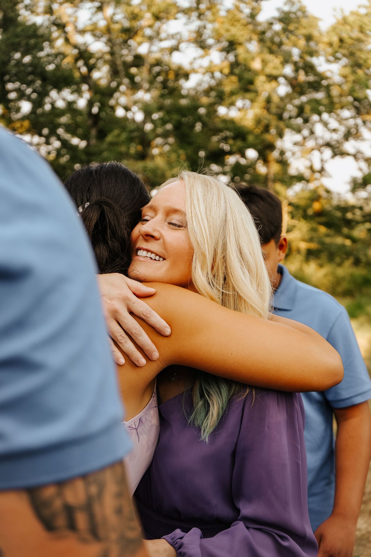 Smiling blonde woman in purple dress joyfully hugging someone outdoors at golden hour.