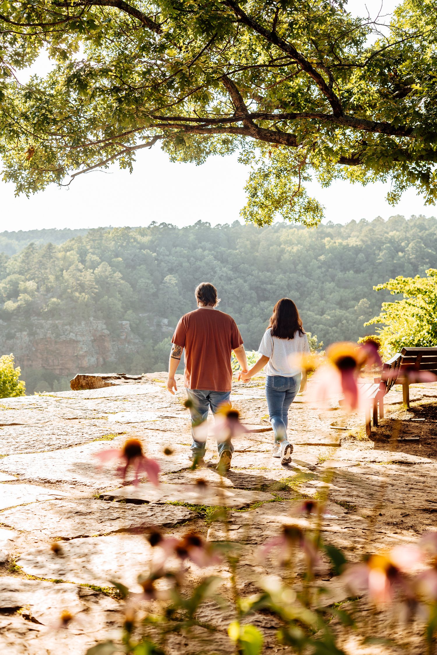 Couple holds hands walking toward a scenic overlook with wildflowers and lush green forest views.