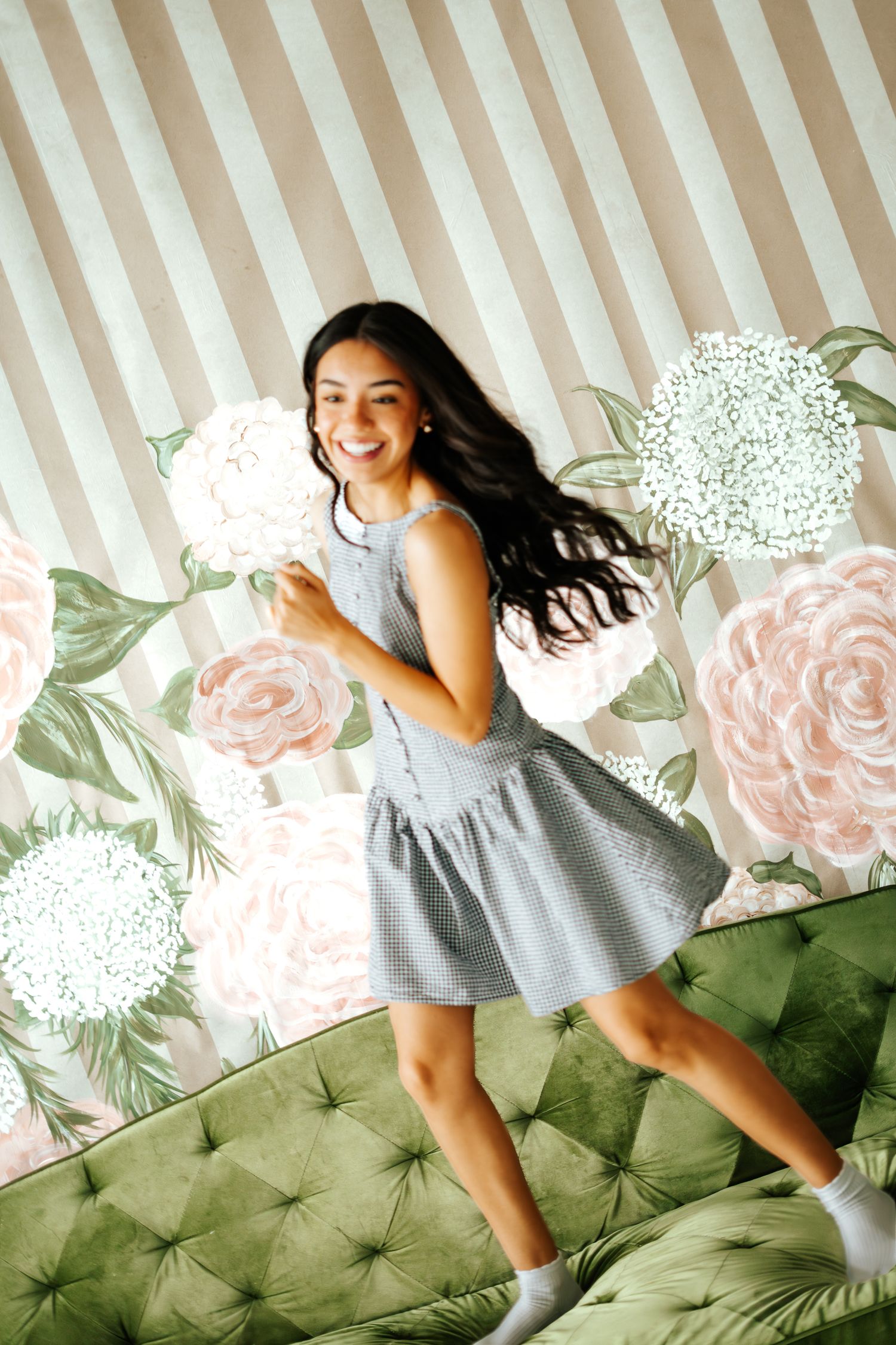 Joyful young woman in a gray dress jumping on a green velvet sofa with floral wallpaper behind her.
