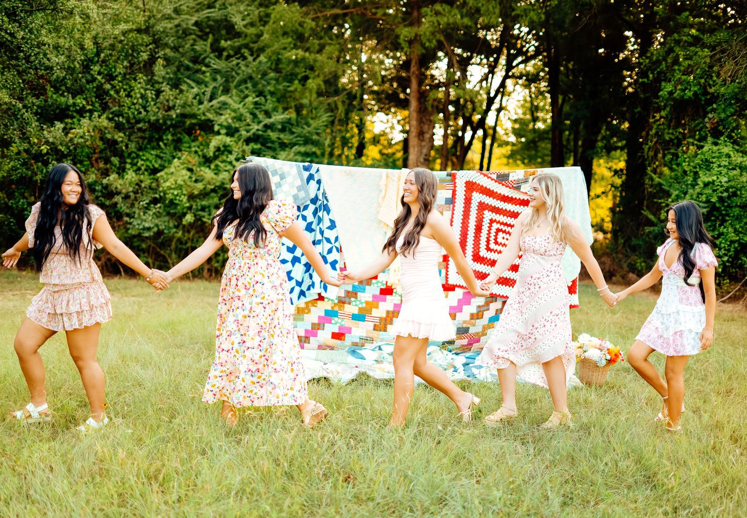 Five young women in floral dresses holding hands and dancing in a green field with quilts.