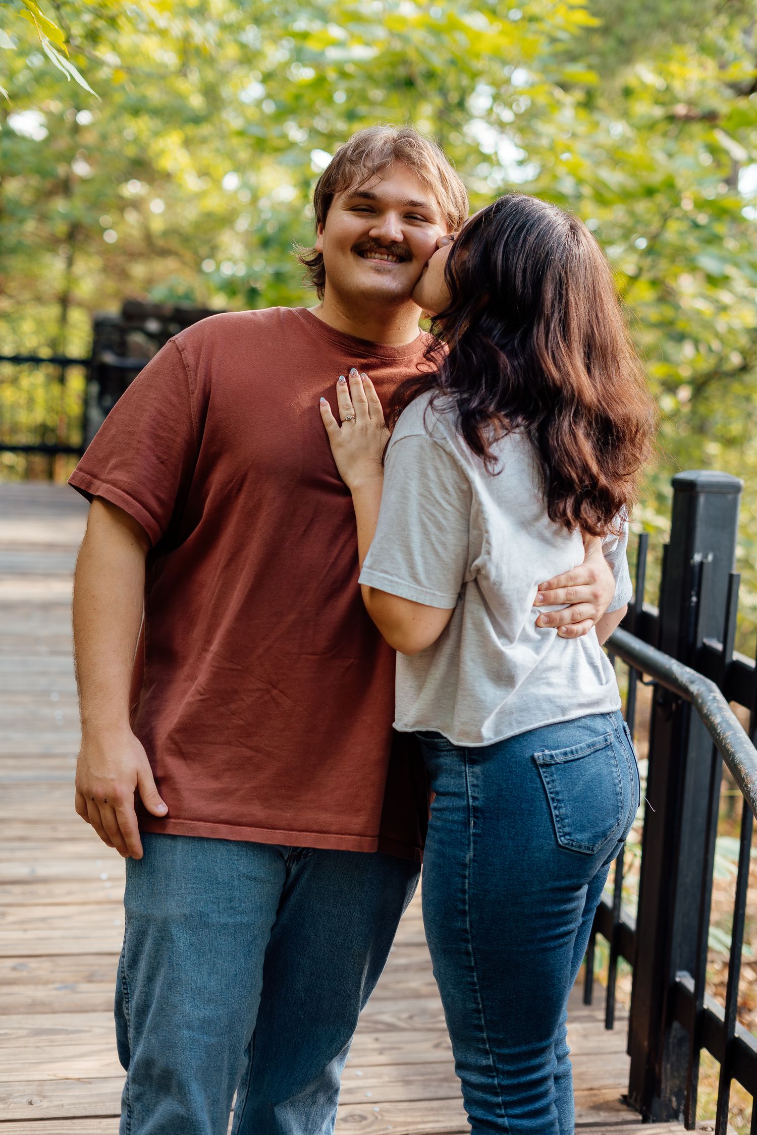 Happy couple shares a tender kiss on a wooden bridge surrounded by green trees in an outdoor park.
