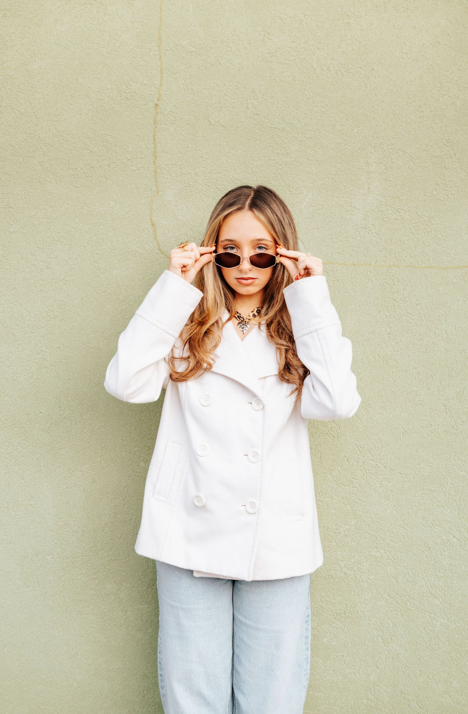 Stylish young woman in white coat adjusting sunglasses while standing against a sage green wall.
