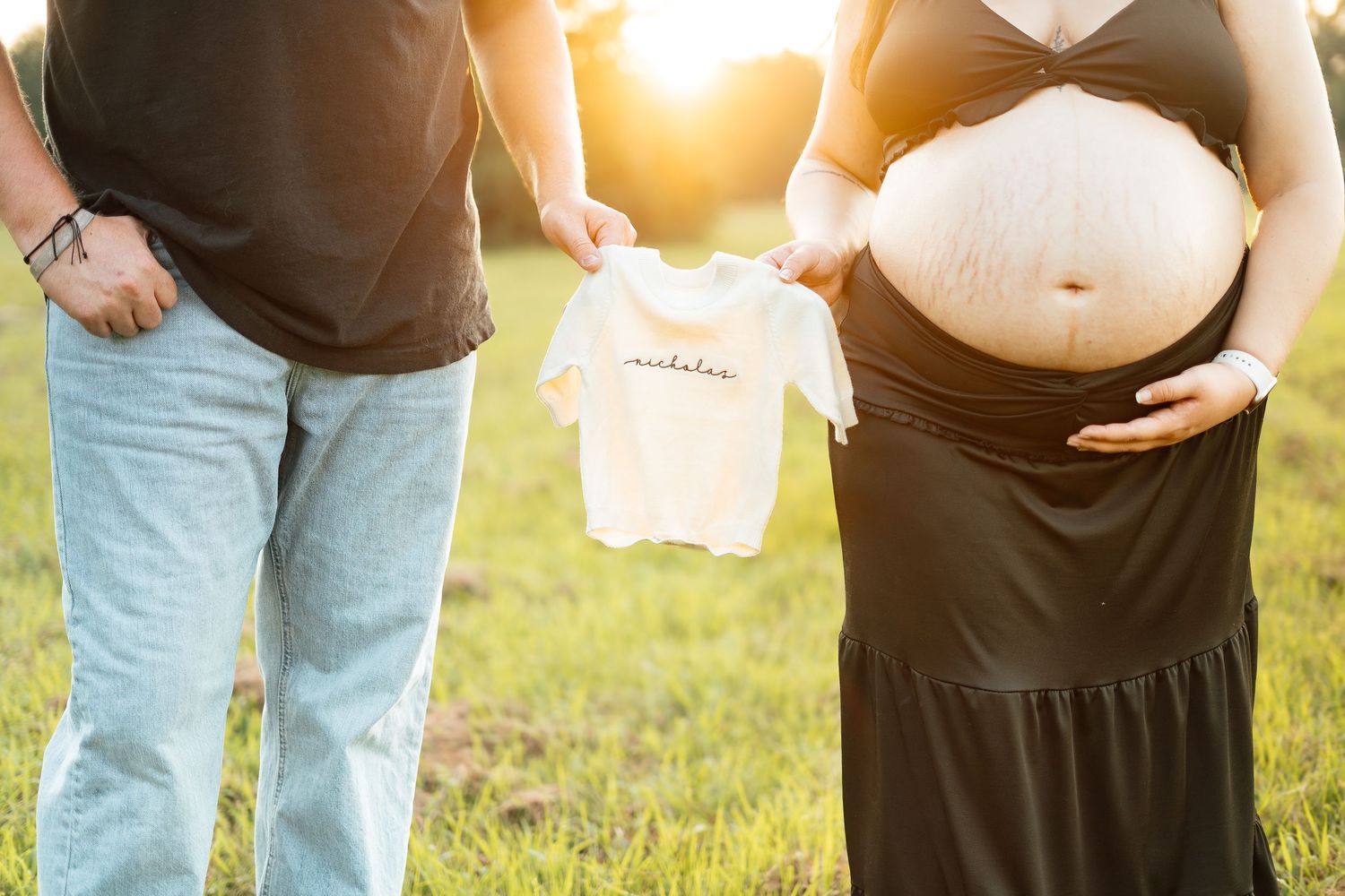 Couple holding tiny baby onesie outdoors during maternity photoshoot at golden hour.