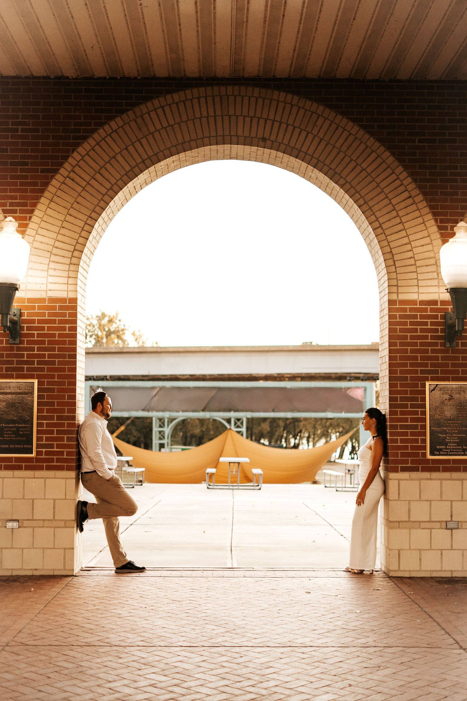 Bride and groom standing apart under a brick archway with warm golden sunlight glowing behind them.